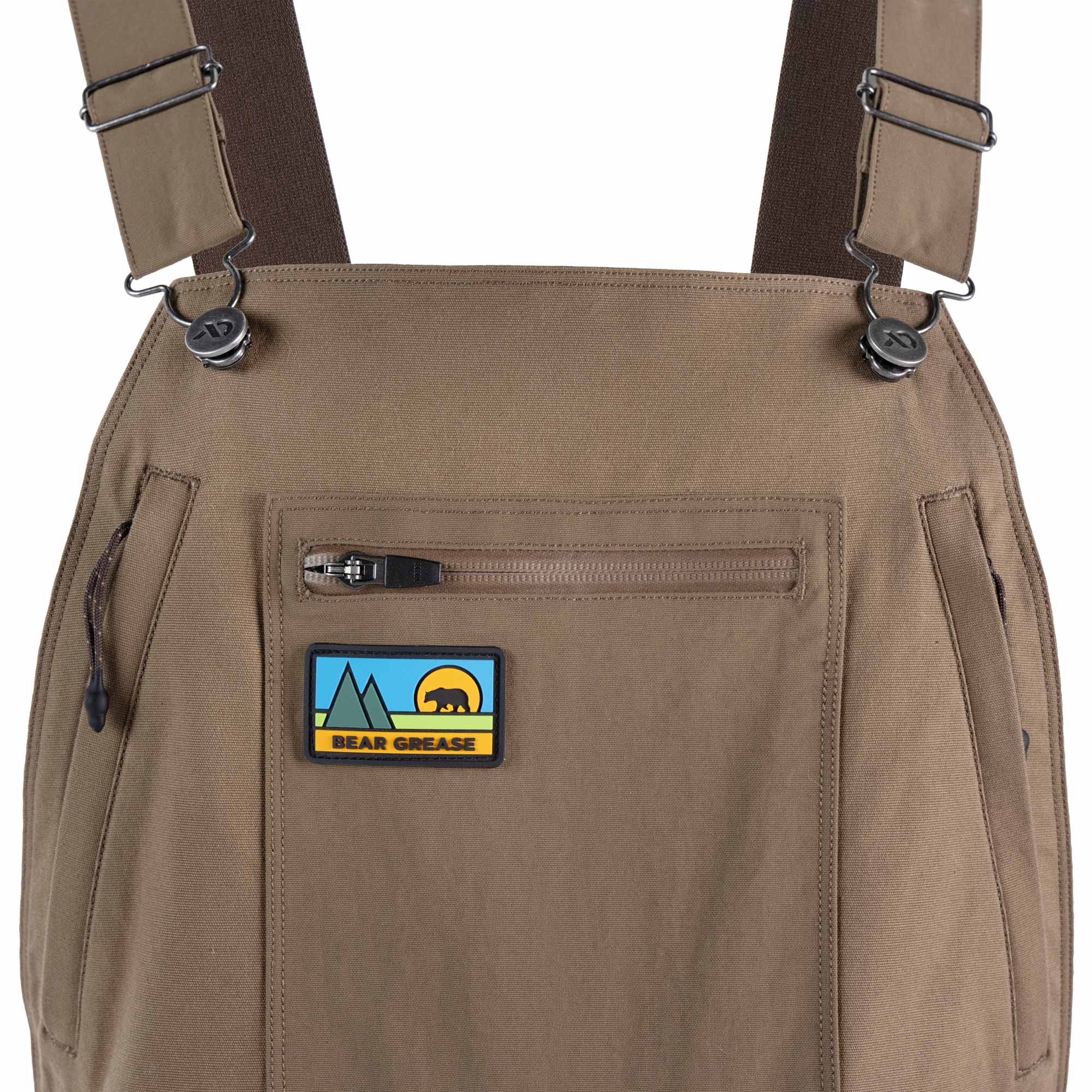 Close-up of tan bib overalls with adjustable shoulder straps, side pockets, and a front zip pocket featuring a 'Bear Grease' patch.