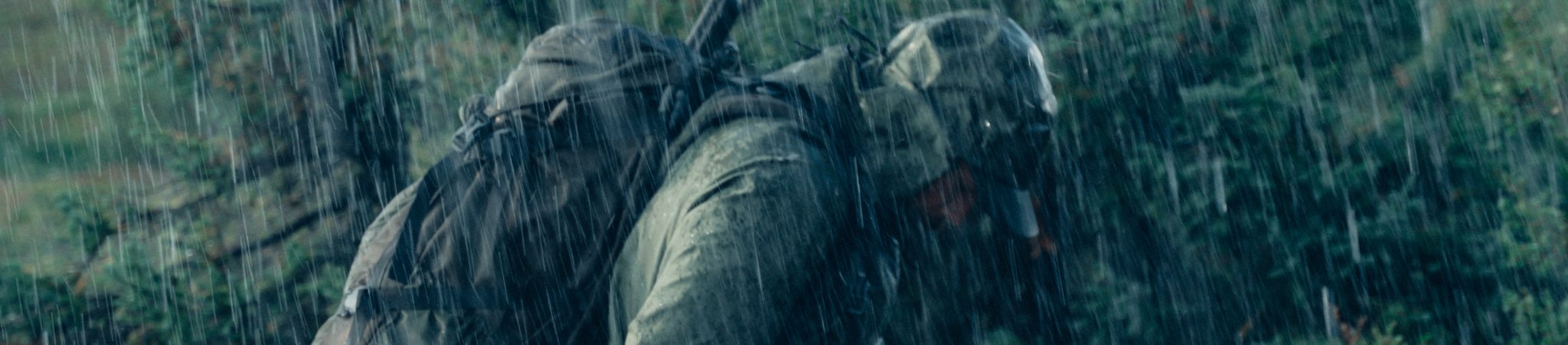 A person wearing a dark raincoat and a hood, crouching in heavy rain outdoors against a blurred forest background.
