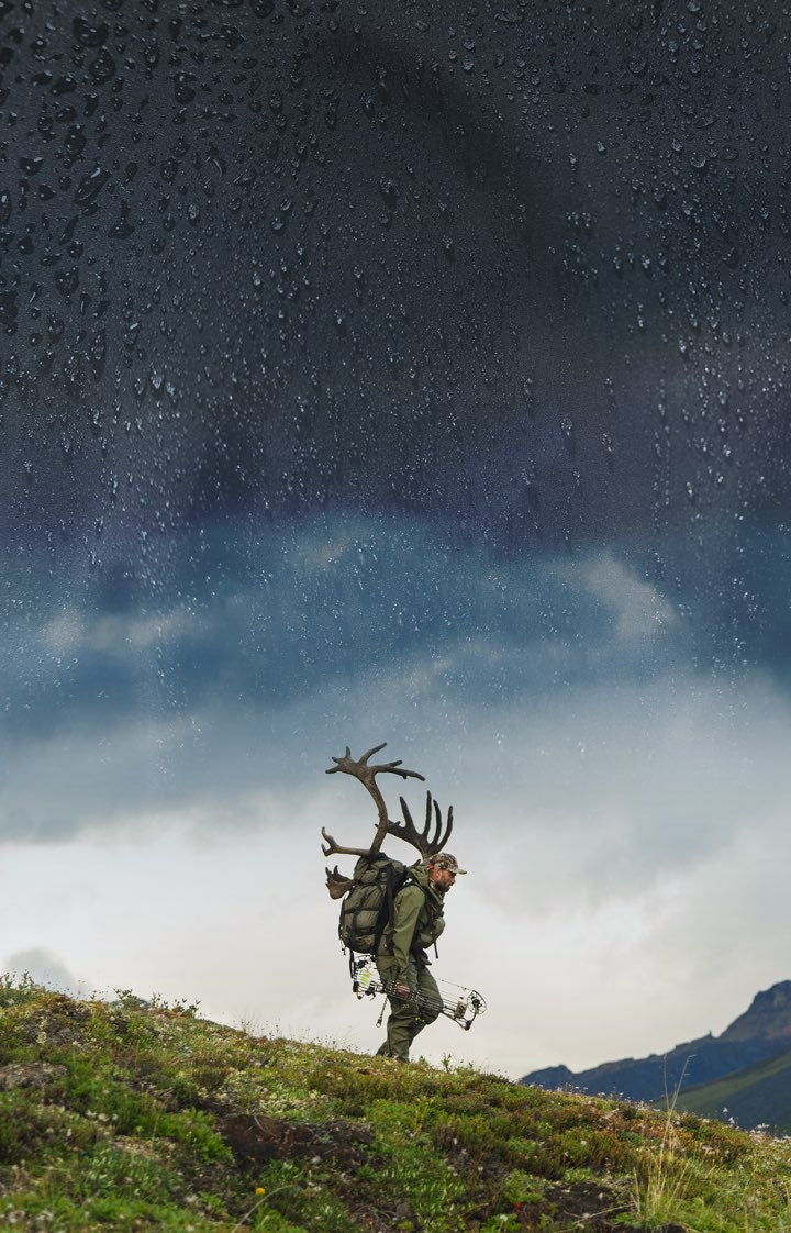 A lone hunter in rain gear walks along a grassy ridge carrying a large antlered caribou on their back, set against a dramatic, rain-splattered dark sky.