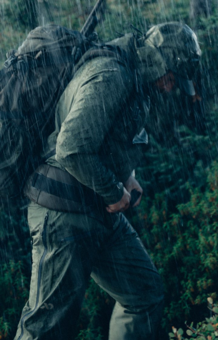 A person in a hunting rain suit with hood carries a large backpack while walking through dense foliage in heavy rain.
