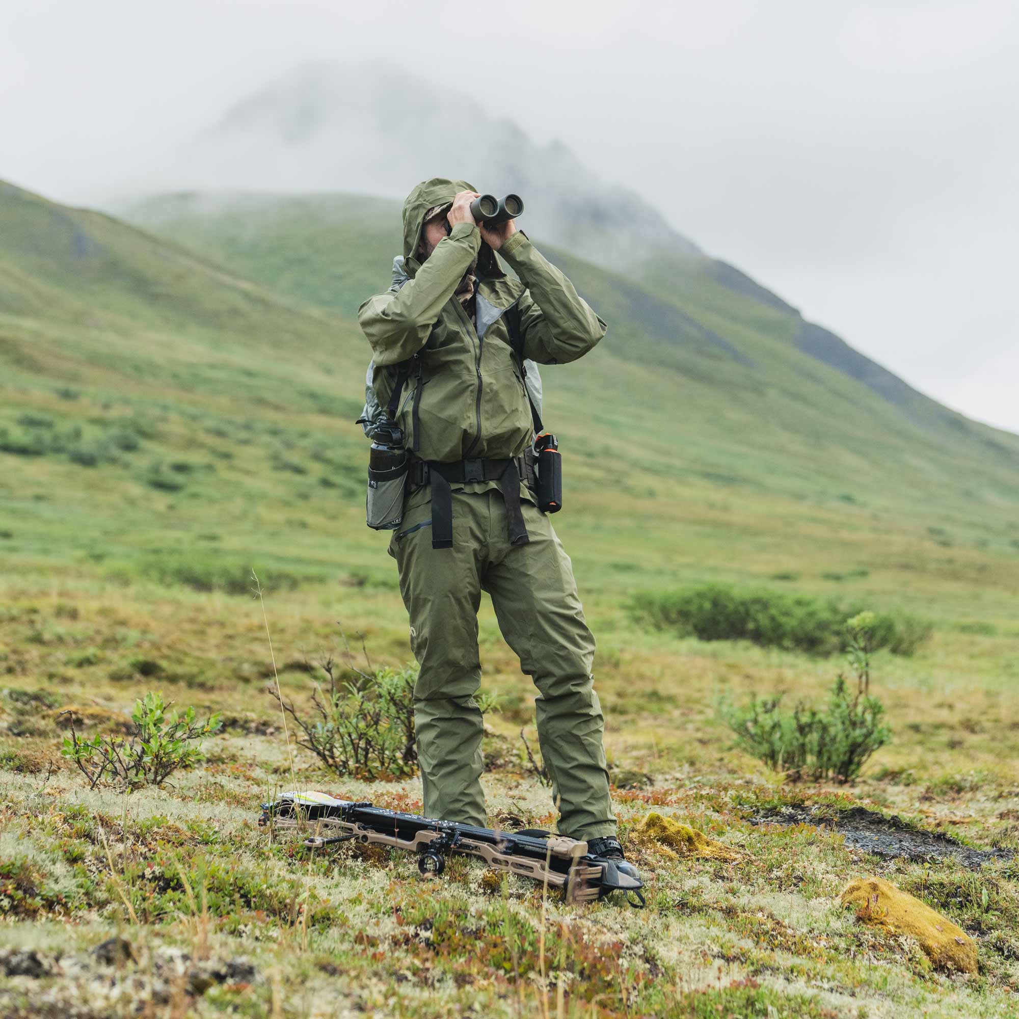 A person in an olive green outdoor hunting suit stands on a grassy mountain slope using binoculars, with a rifle resting on the ground in front of them and misty hills in the background.