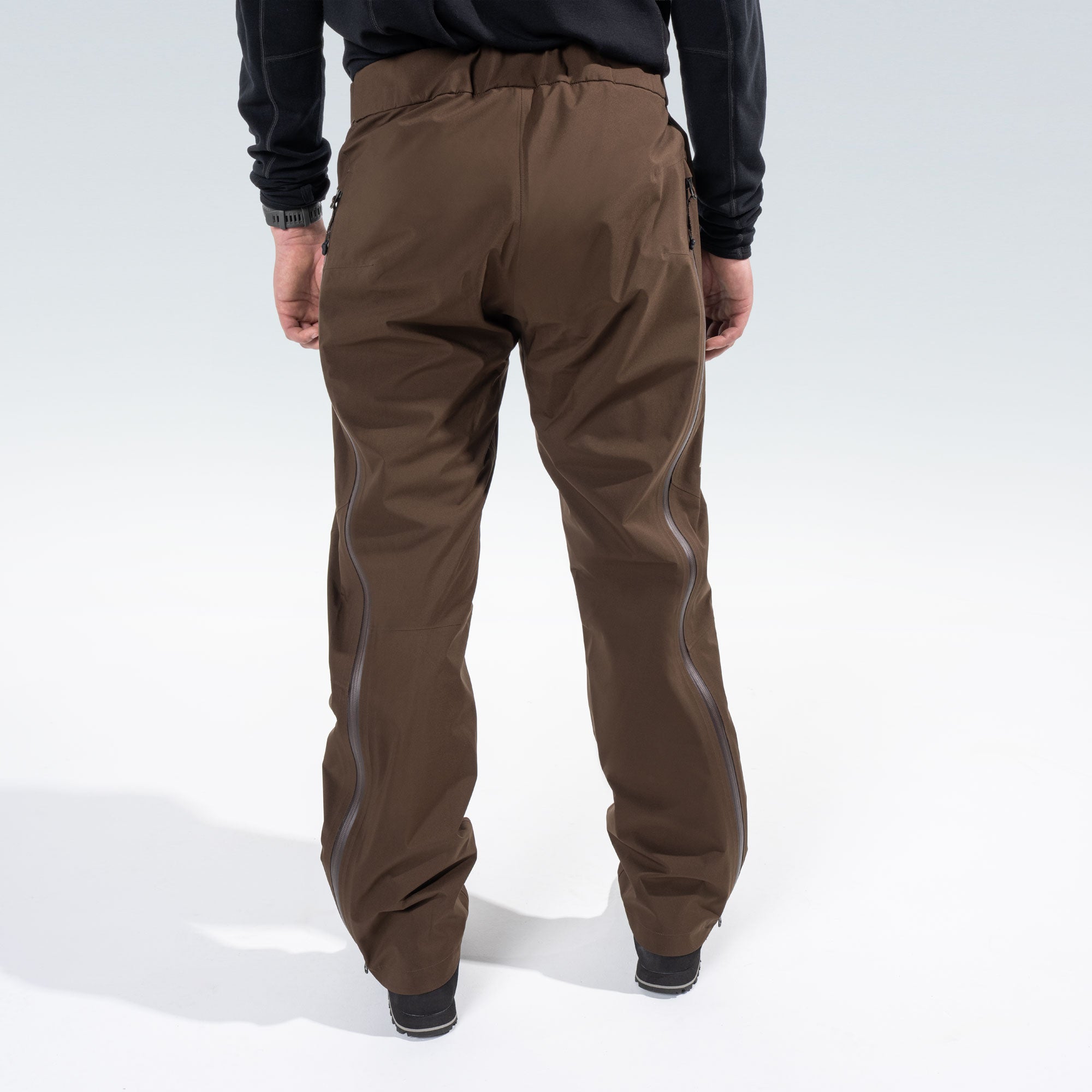 Rear view of a person wearing brown technical pants with side zips, paired with a black long-sleeve top, against a light gray studio background.