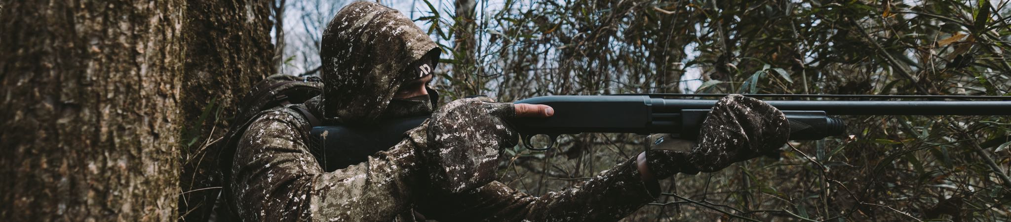 Camouflage-clad person in a hooded jacket and gloves aims a black shotgun.