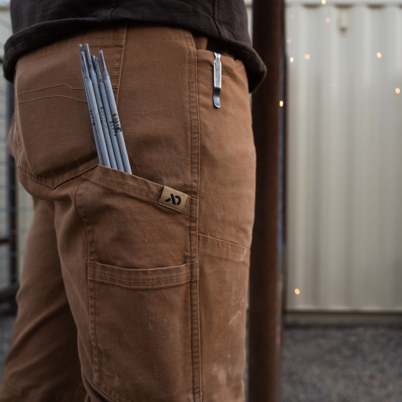 Close-up of a person wearing brown canvas utility pants with side pockets, photographed outdoors against a light corrugated wall.