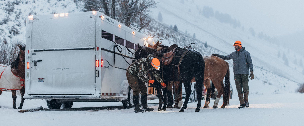 Two hunters in heavy jackets and cold weather gear place saddles on a group of horses tethered to a trailer.