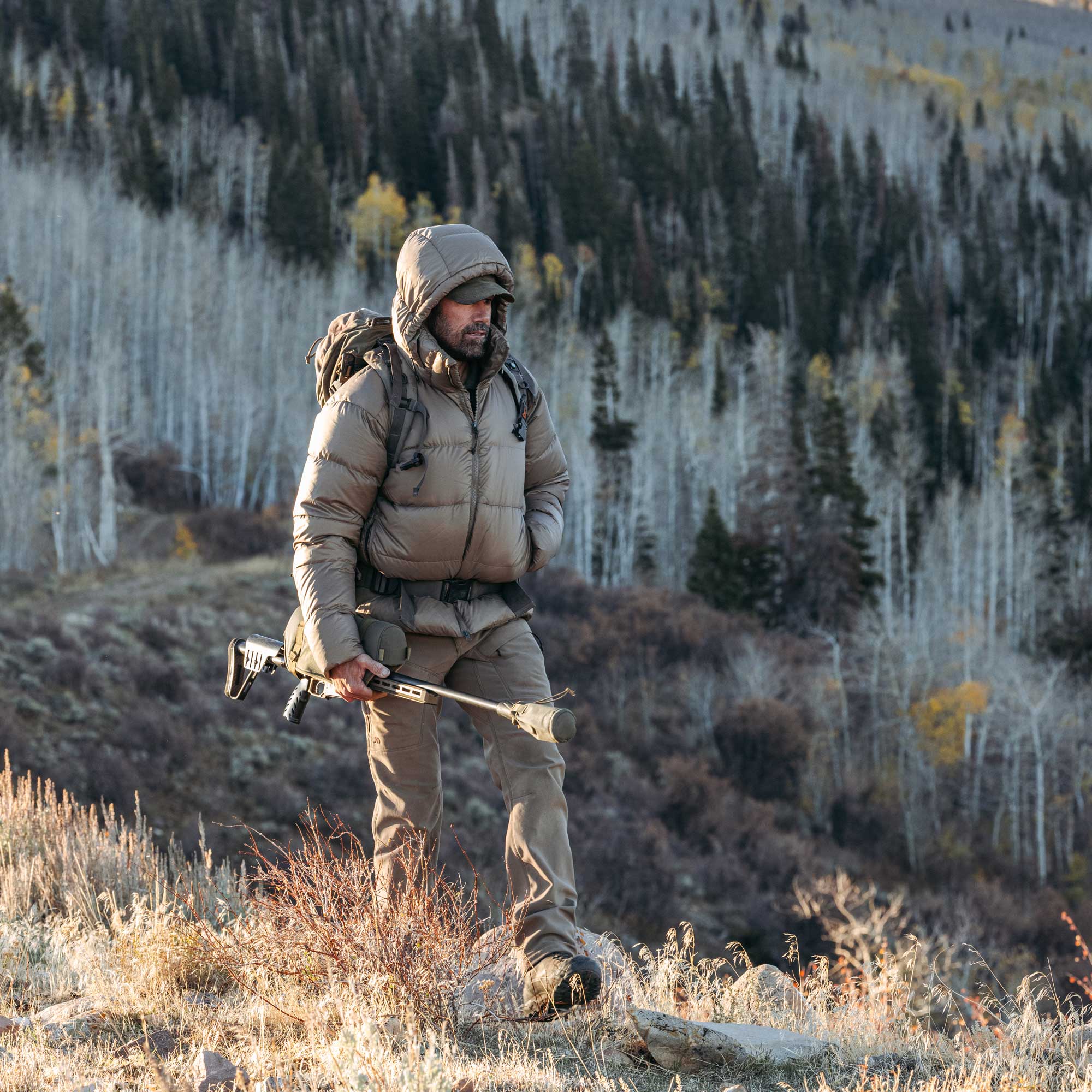 A man in a beige puffer jacket and hiking backpack stands on a grassy mountainside holding a rifle, with a forested hillside in the background.