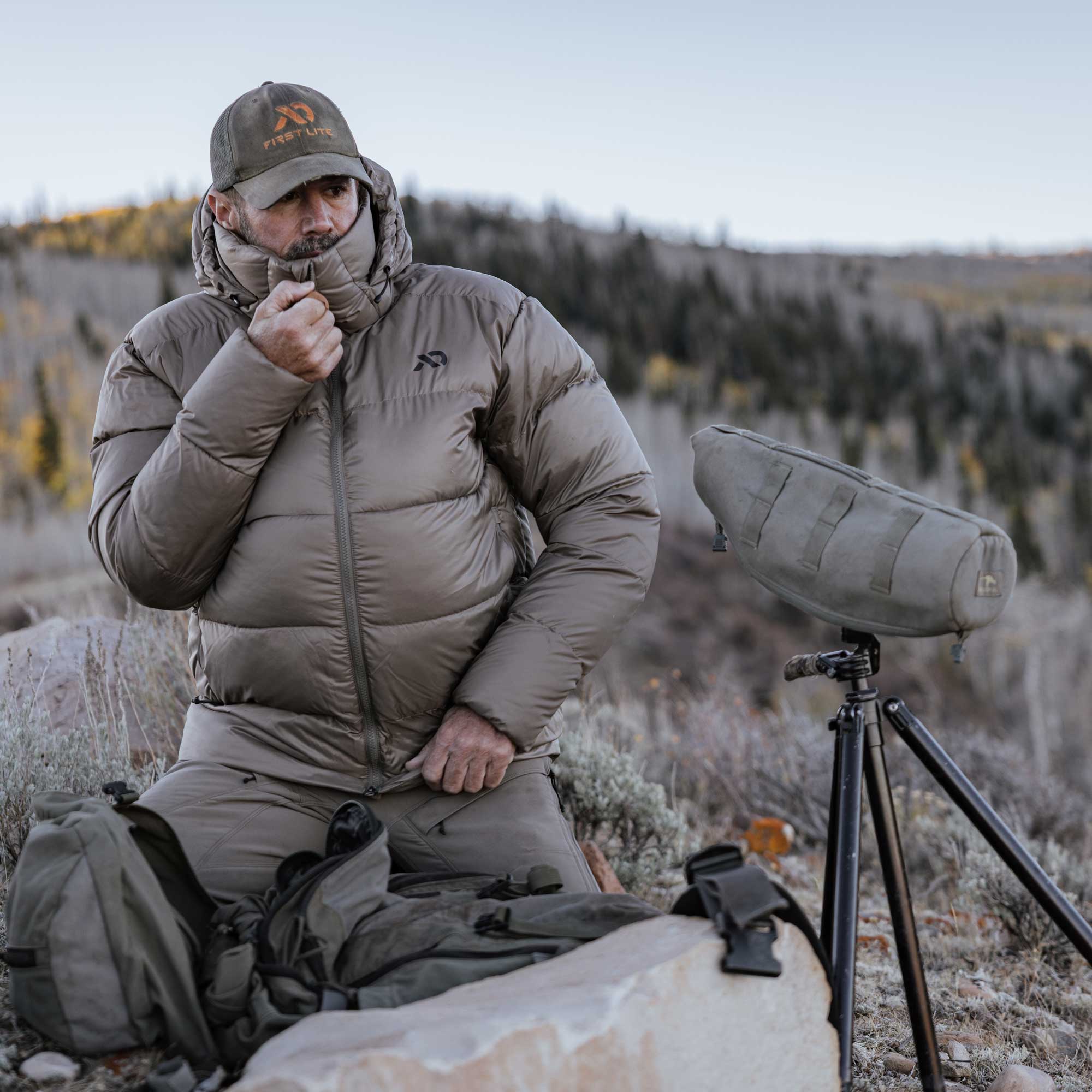 A man in outdoor hunting attire sits on a rock in a mountain setting, wearing a taupe insulated puffer jacket, matching pants, and a cap with a visible First Lite logo. A large spotting scope on a tripod and backpacks are placed beside him.