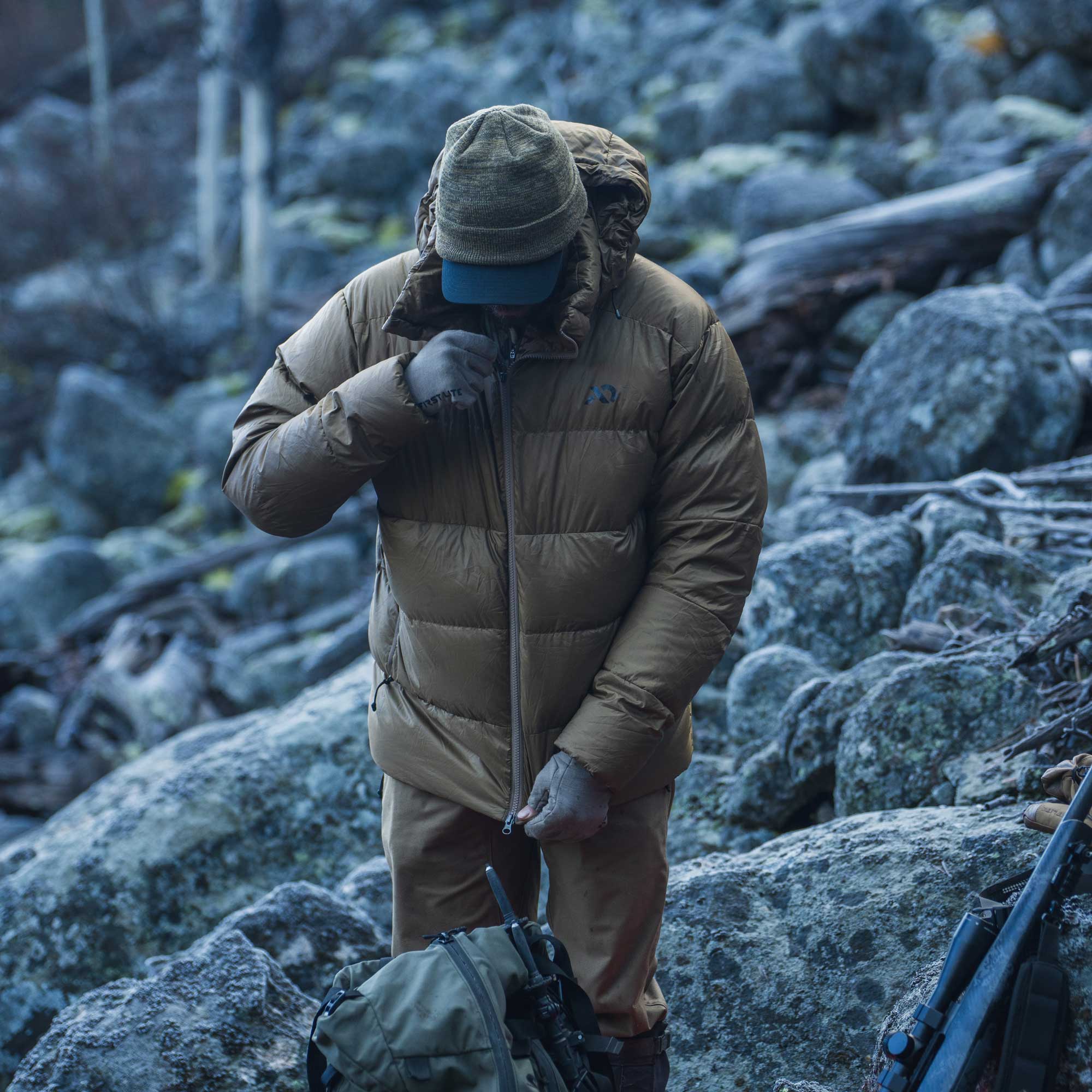A person standing on a rocky outdoor terrain wearing a tan insulated puffer jacket, matching pants, gloves, and a cap, with a backpack in the foreground.