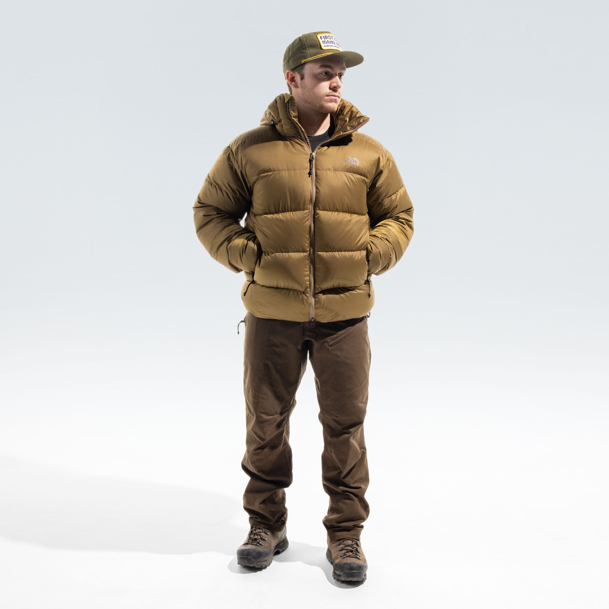 A man wearing a brown puffer jacket, brown pants, and an olive green cap, standing against a light gray studio background.