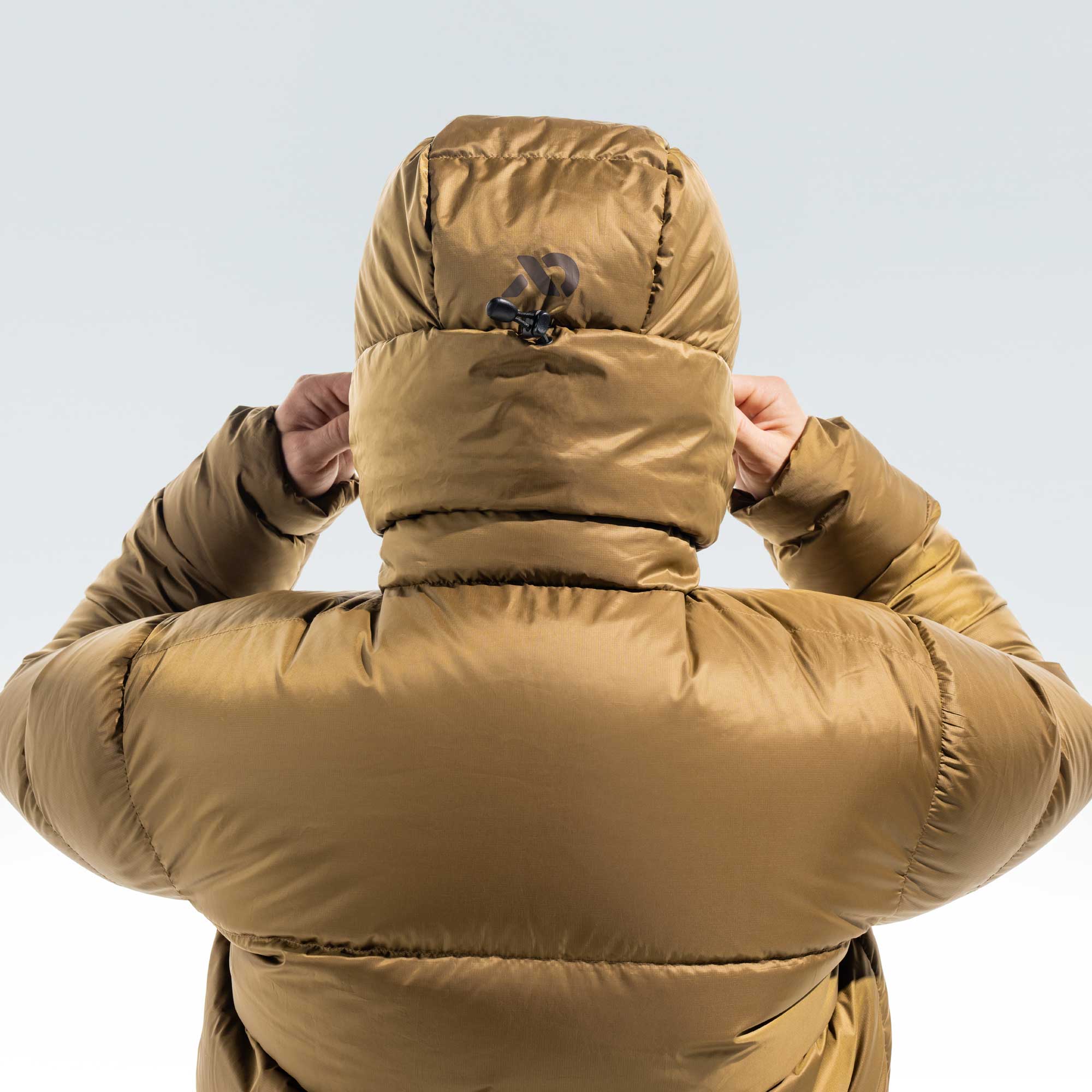 A person viewed from behind wearing a thick, puffy hooded puffer jacket in a warm camel-brown color, adjusting the hood against a light gray studio background.