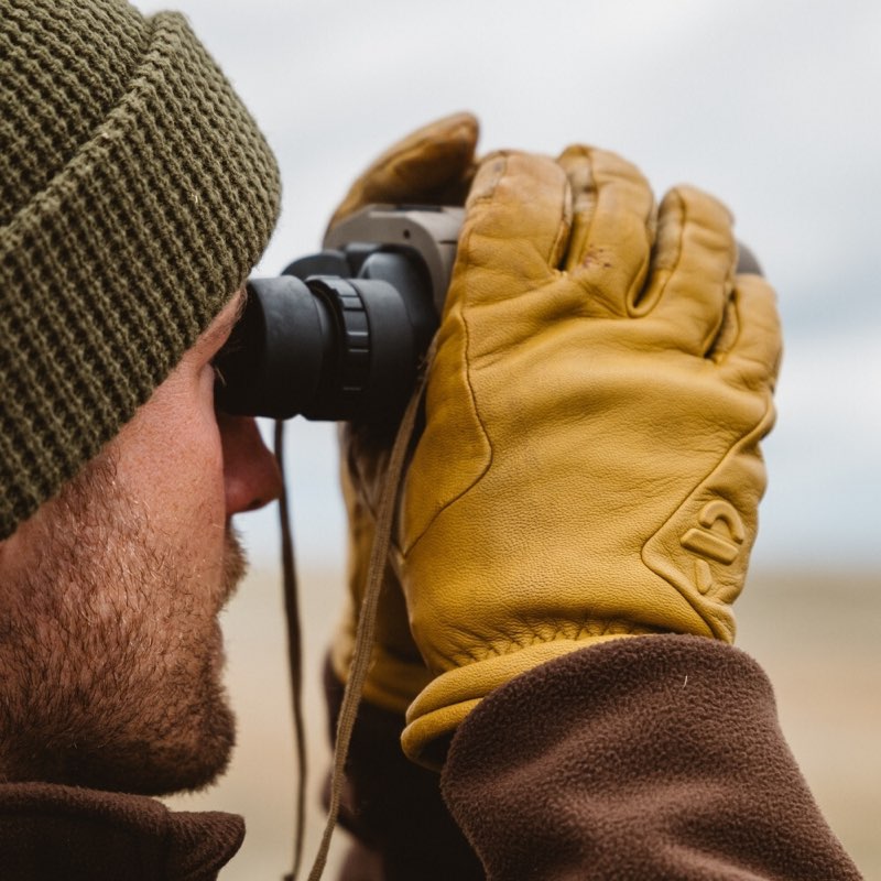 A close-up lifestyle shot of a person wearing a green knit beanie, brown fleece jacket, and a thick yellow leather hunting glove while looking through black binoculars.
