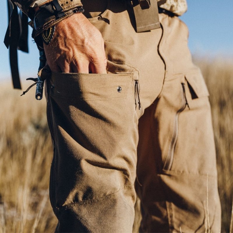 Close-up of a person wearing tan tactical cargo pants with a hand in the front pocket, outdoors in a grassy field under bright sunlight.
