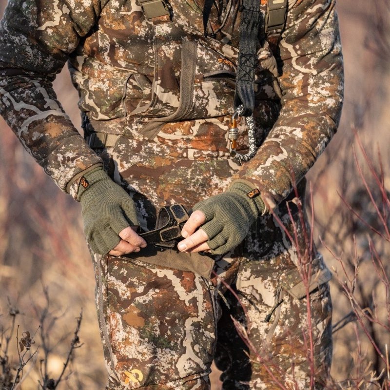 A person wearing a full camouflage hunting suit and matching fingerless gloves, standing outdoors in dry brush while holding a black pouch at the waist.