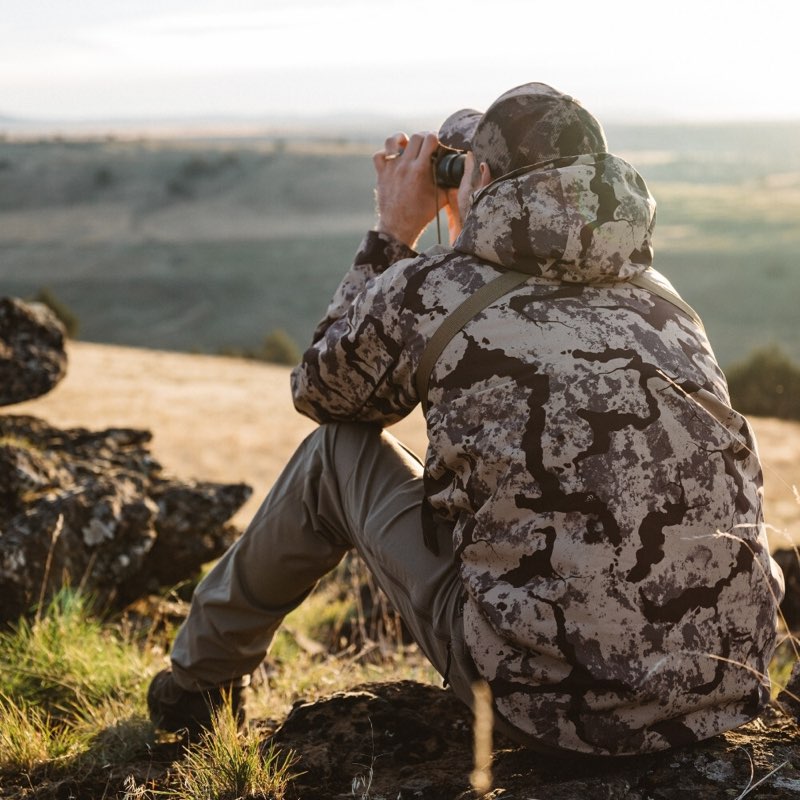 A person wearing a camouflage hunting jacket and pants sits on a rocky hillside, looking through binoculars toward a distant landscape at golden hour.