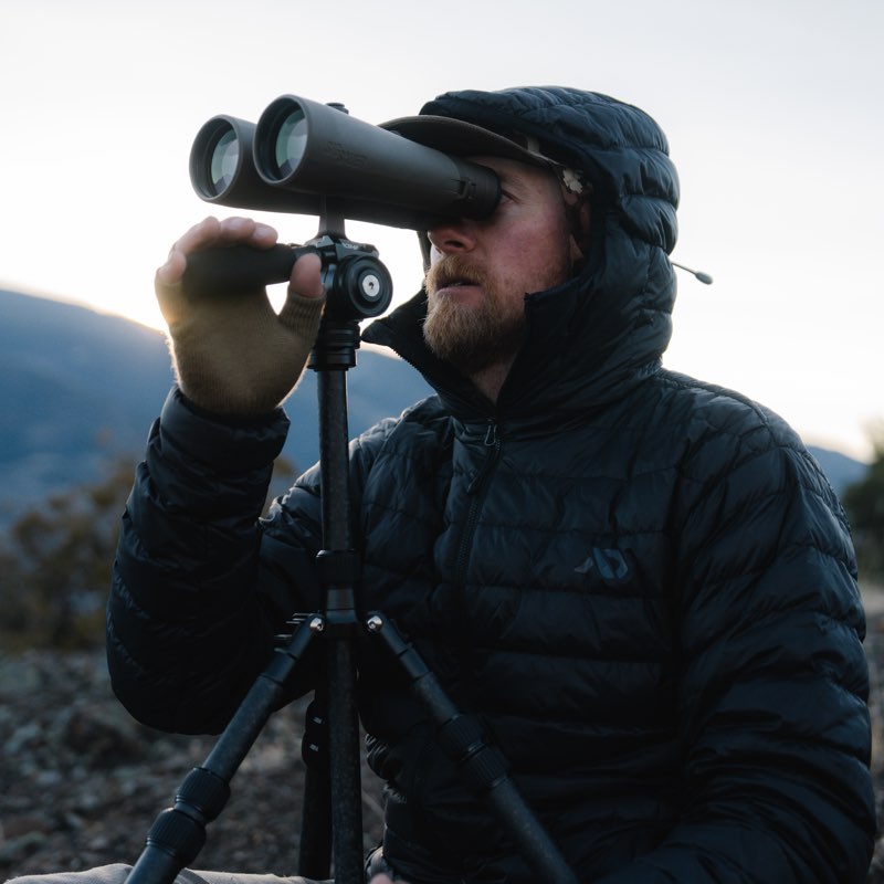 A man wearing a black puffer jacket and cap uses binoculars mounted on a tripod outdoors against a soft mountain background.