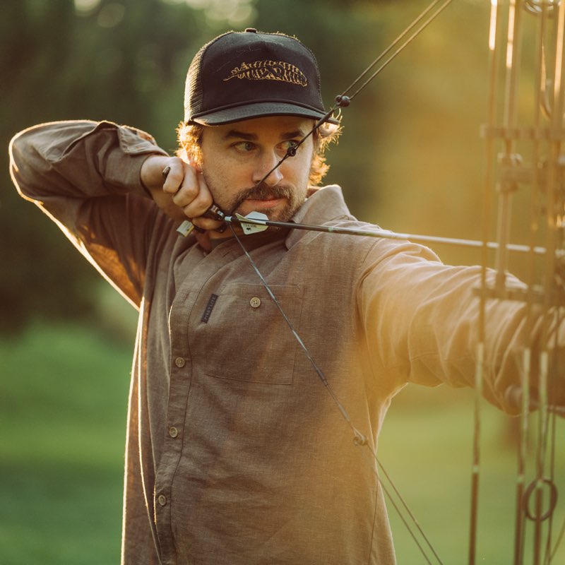 A man in a brown button-up shirt and a black baseball cap with gold lettering draws a bow in an outdoor setting with warm golden-hour sunlight.