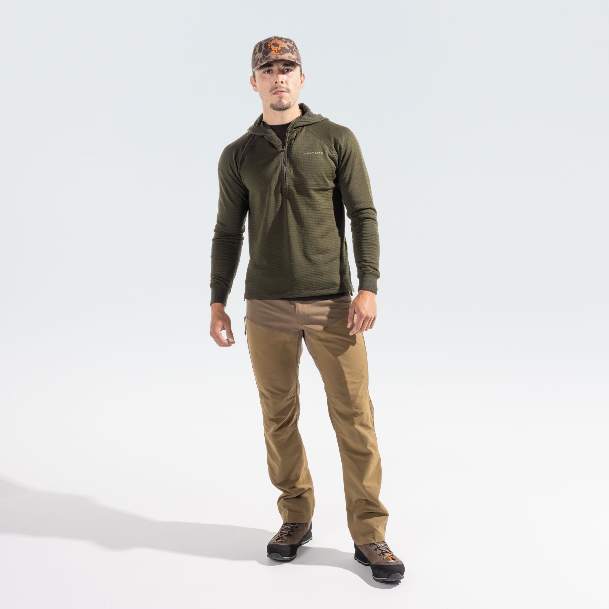 A man modeling an olive green quarter-zip hooded pullover, tan pants, and a camouflage cap against a light gray studio background.