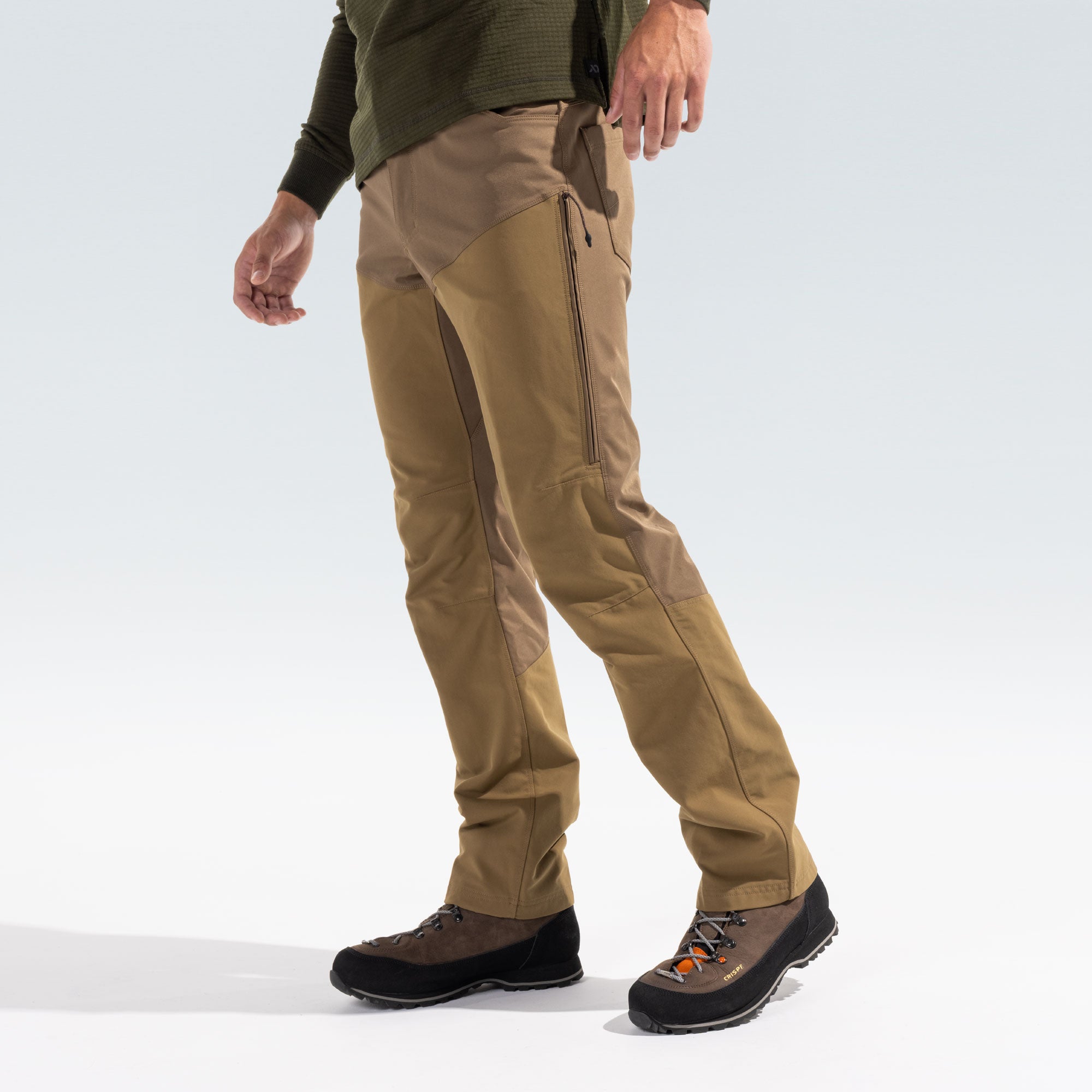 A person wearing tan technical outdoor pants with zippered knee panels and dark hiking boots, shown from the waist down against a light gray studio background.