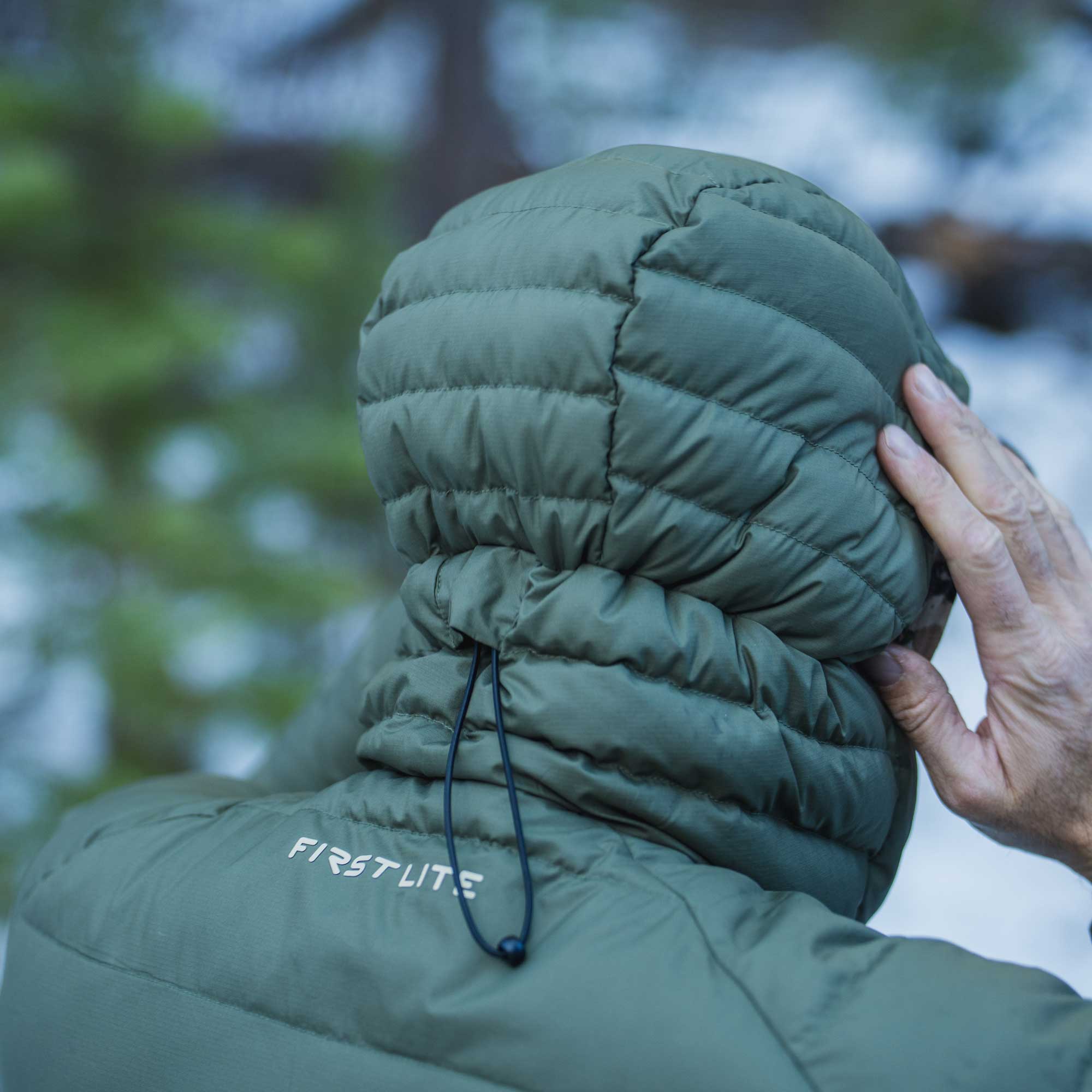 Close-up back view of a person wearing an olive green puffer jacket outdoors, with the hood up and a hand adjusting it. The jacket has a visible FIRSTLITE logo on the shoulder.