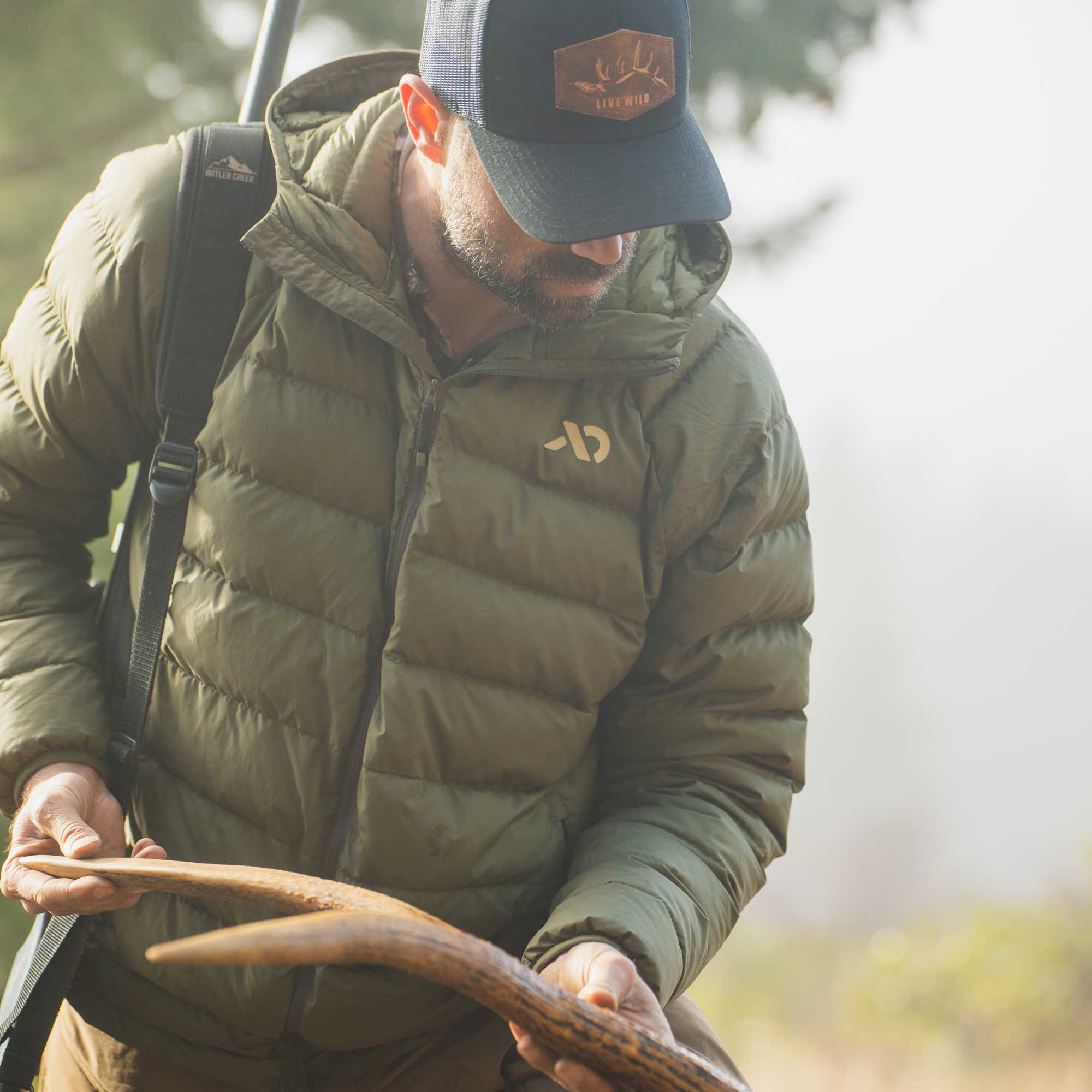 A man in outdoor hunting attire wearing an olive green puffer jacket with a gold logo on the chest, a dark navy cap with a deer head patch, and holding a large antler in his hands.