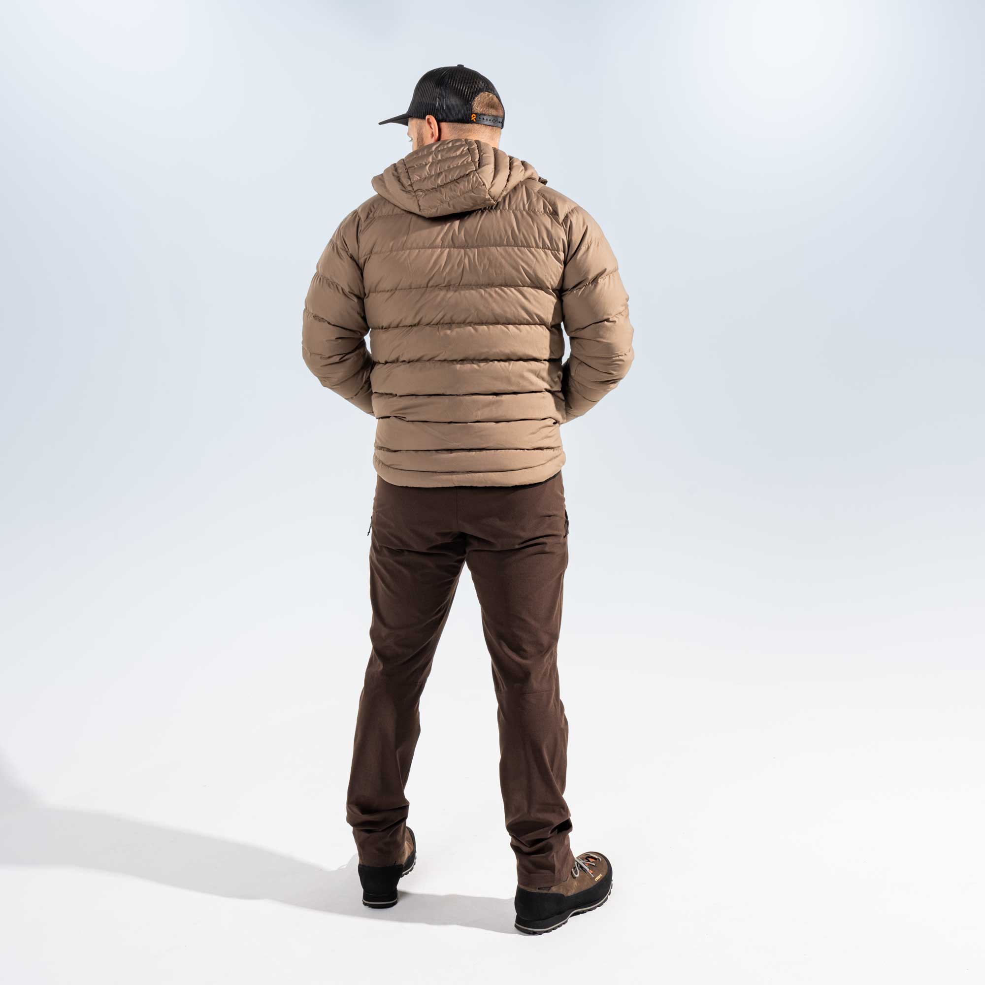 A man viewed from behind wearing a tan hooded puffer jacket, dark brown pants, and a black baseball cap against a light gray studio background.