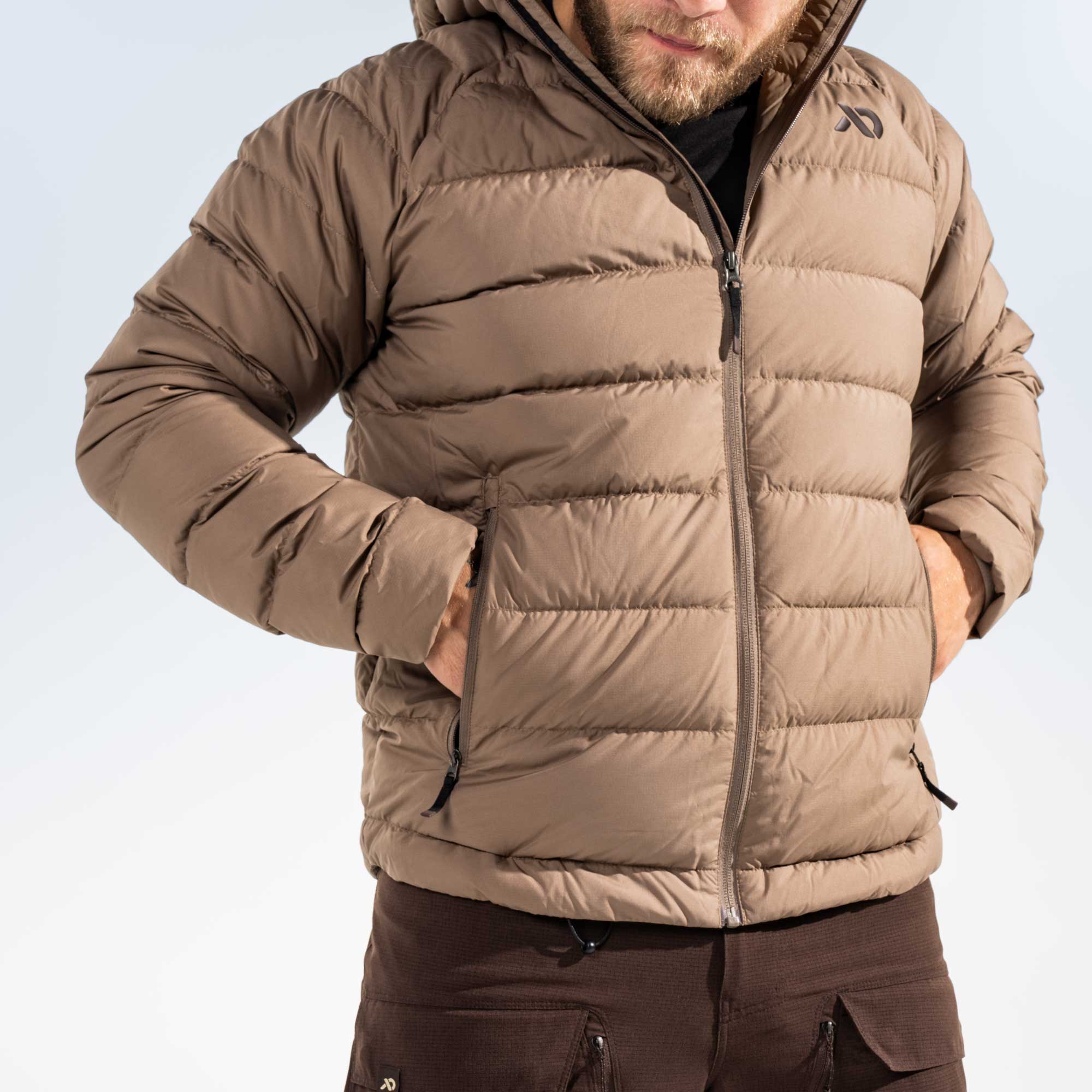 A man wearing a brown hooded puffer jacket with horizontal quilting, shown from the chin down against a light gray studio background.