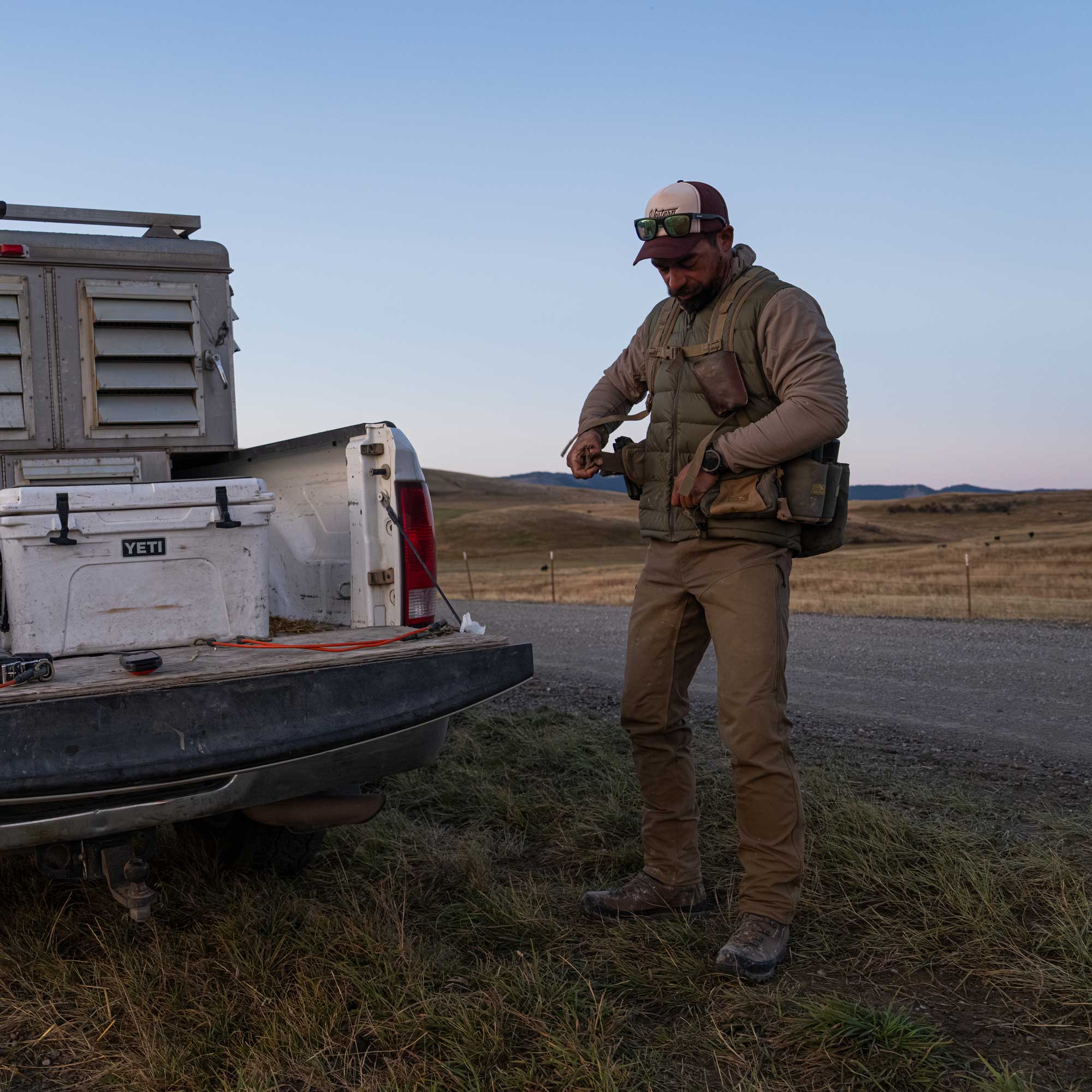 A man standing at a truck with dog boxes in it, putting on an upland hunting vest and wearing a green puffer vest in an upland praire setting.