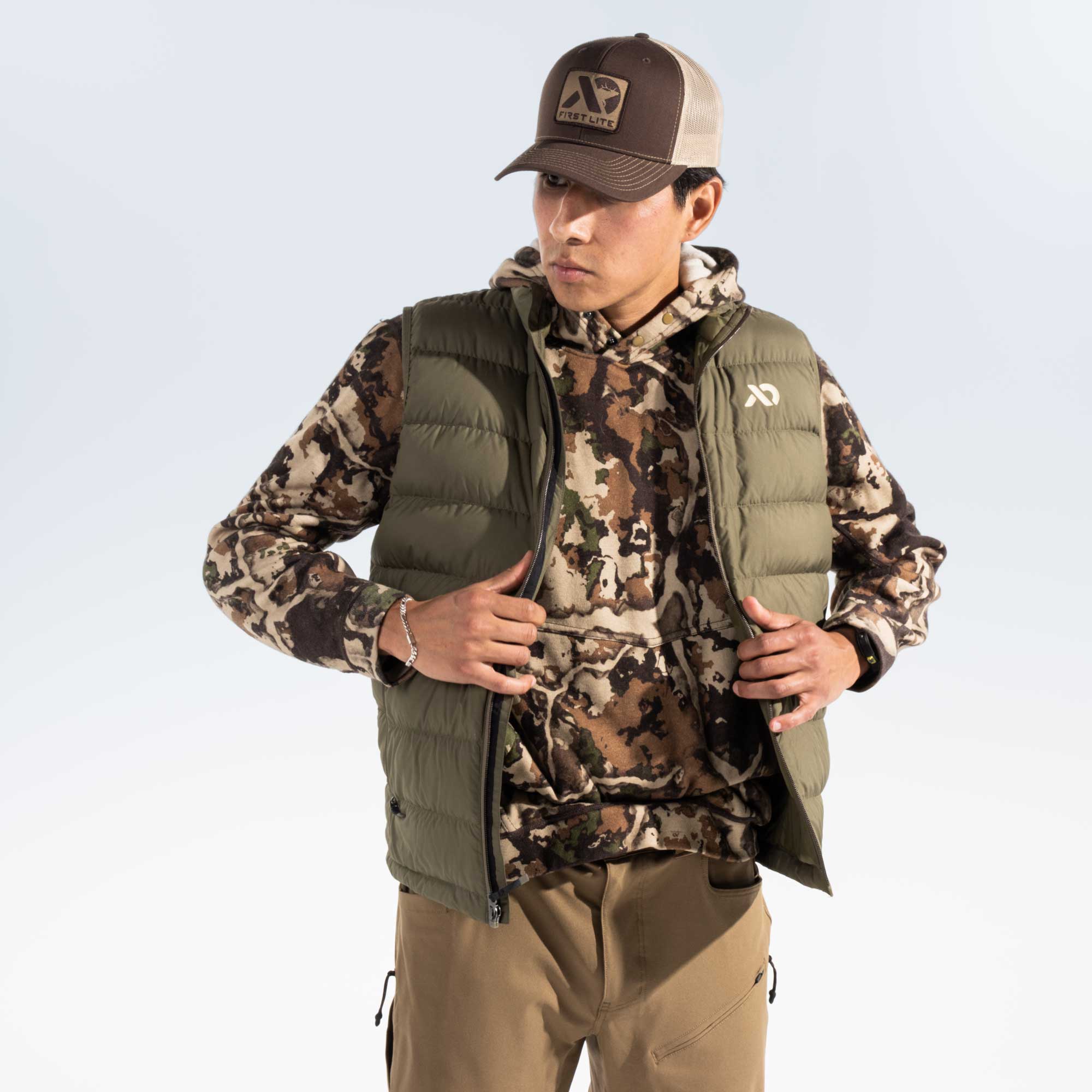 A man wearing a camouflage hoody adjusting an olive green puffer vest against a studio background.