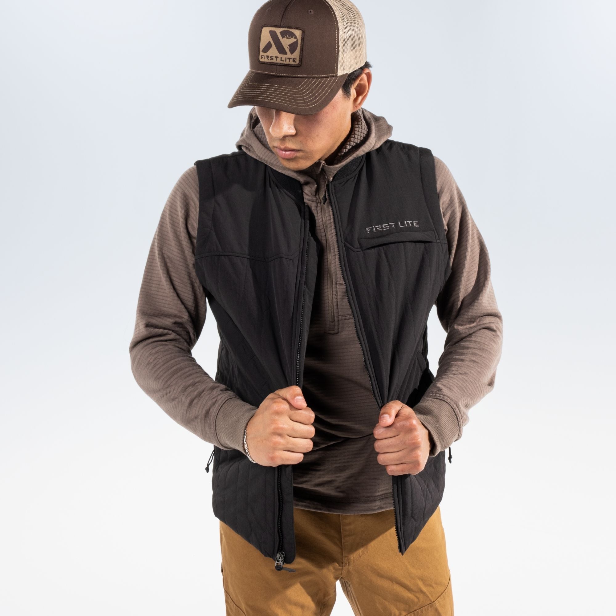 Man wearing a black vest over a brown hoodie, tan pants and a FIRST LITE cap standing against a white studio background.