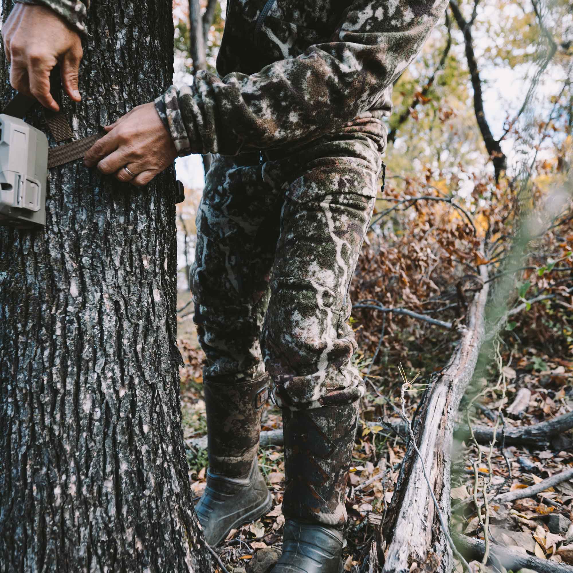 Man wearing camo jacket and pants tying brown straps around a tree.