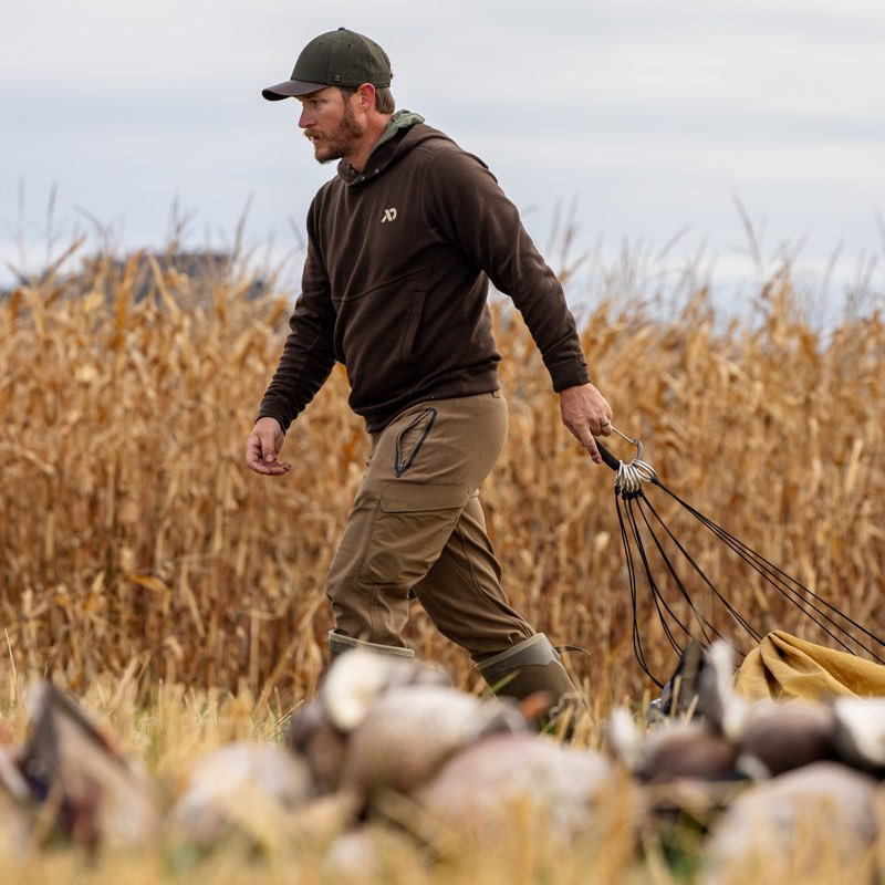 A man wearing a dark brown hoodie, brown pants, and a black cap walks through a field of dry corn while pulling a rope attached to a duck hunting rig. Ducks are visible in the blurred foreground.