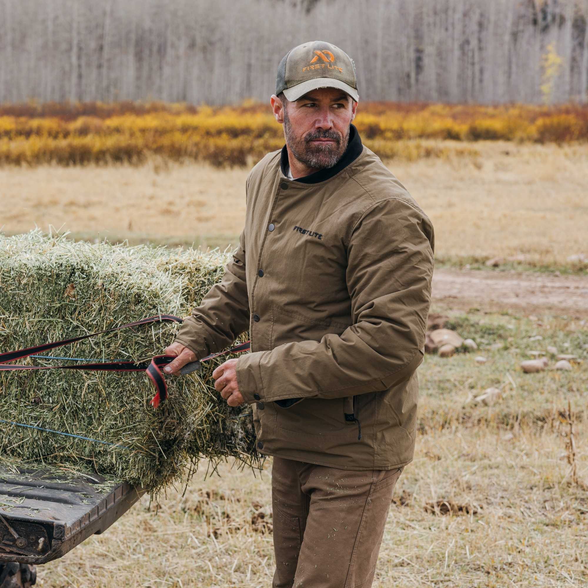 Man wearing an akern chore coat, brown pants and a FIRST LITE hat.