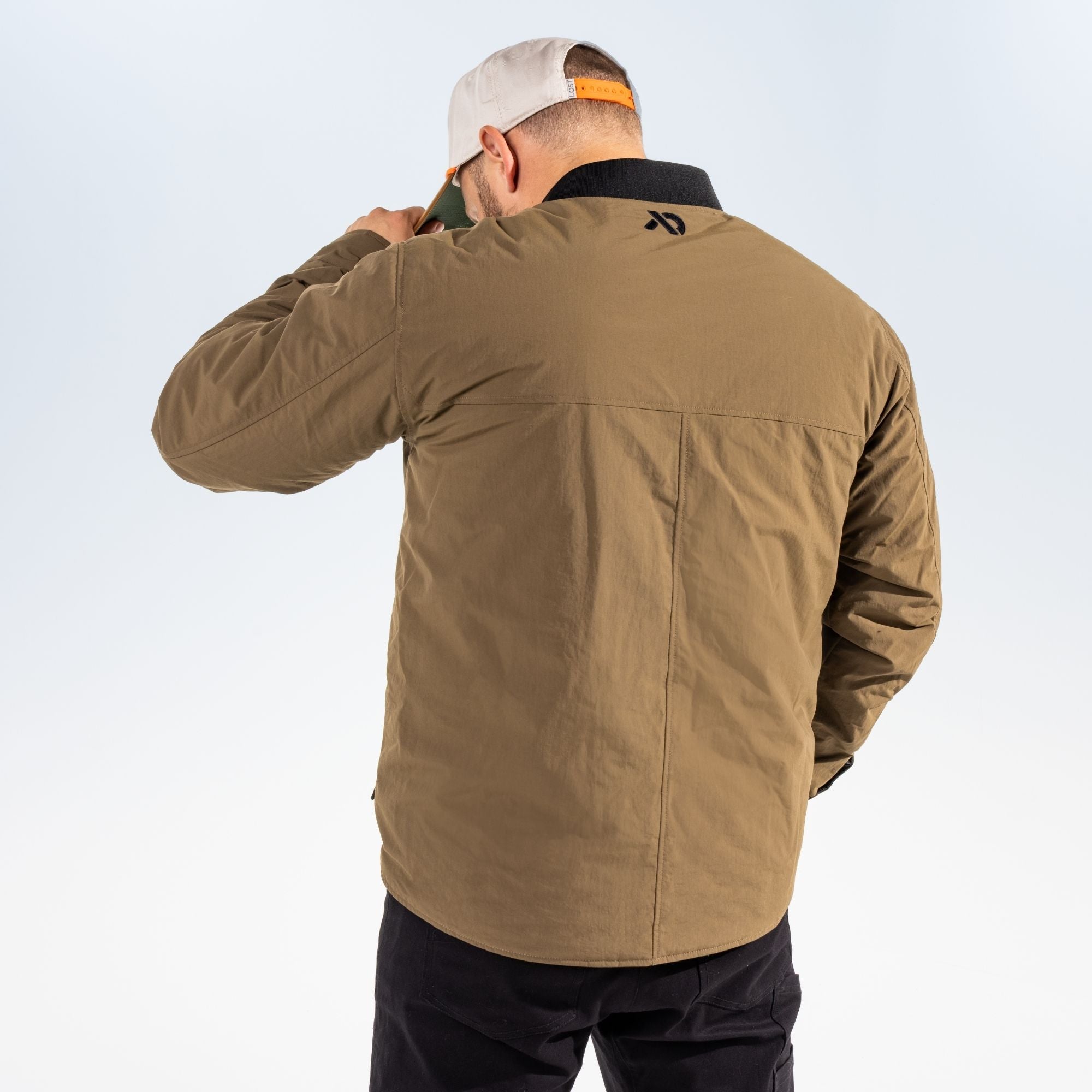 Reverse shot of a man wearing an akern chore coat with a FIRST LITE logo in the upper side of the back and a white hat against a light gray studio background