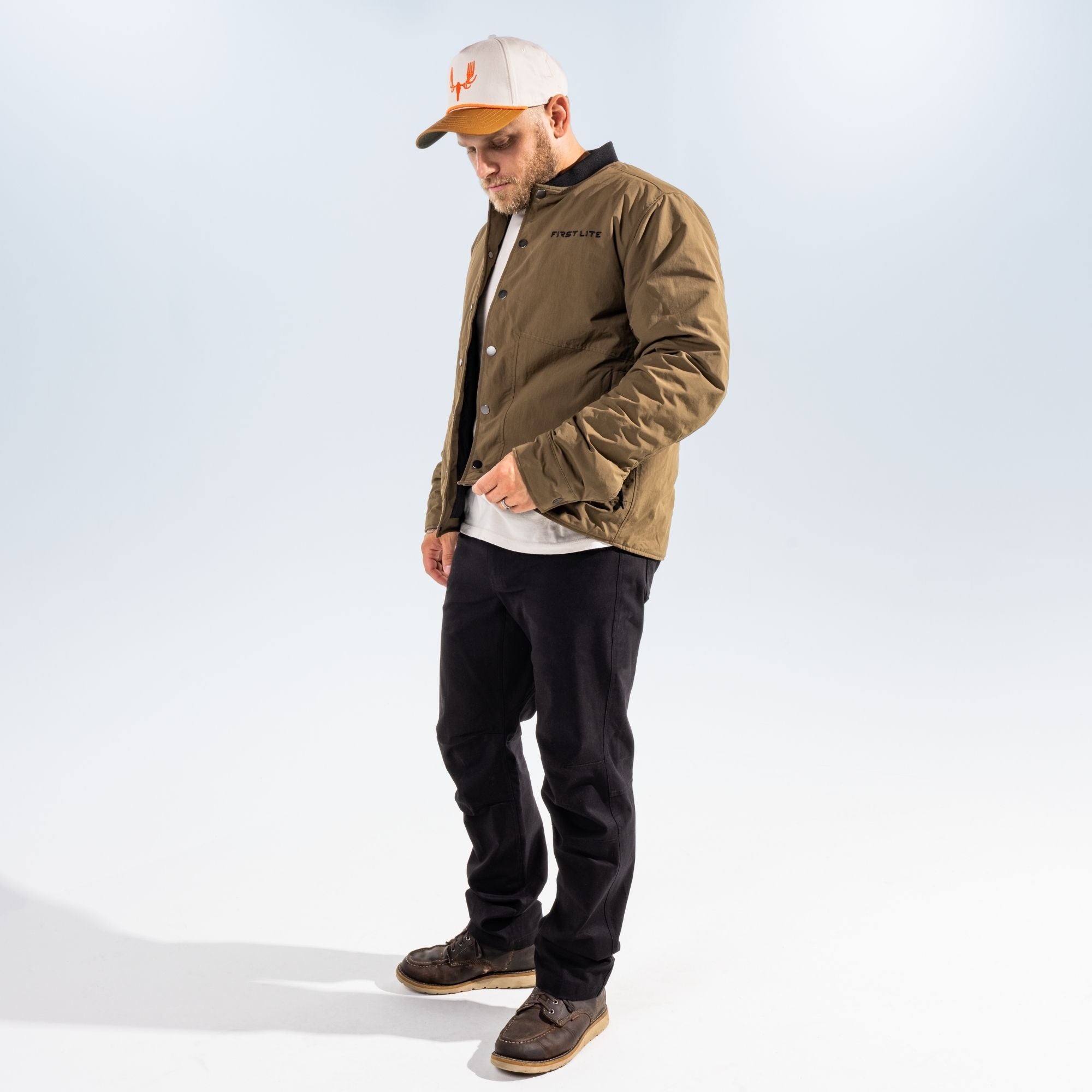 Man wearing an open akern chore coat, a white T-shirt, a hat with MEATEATER logo in orange and black pants against a light gray studio background