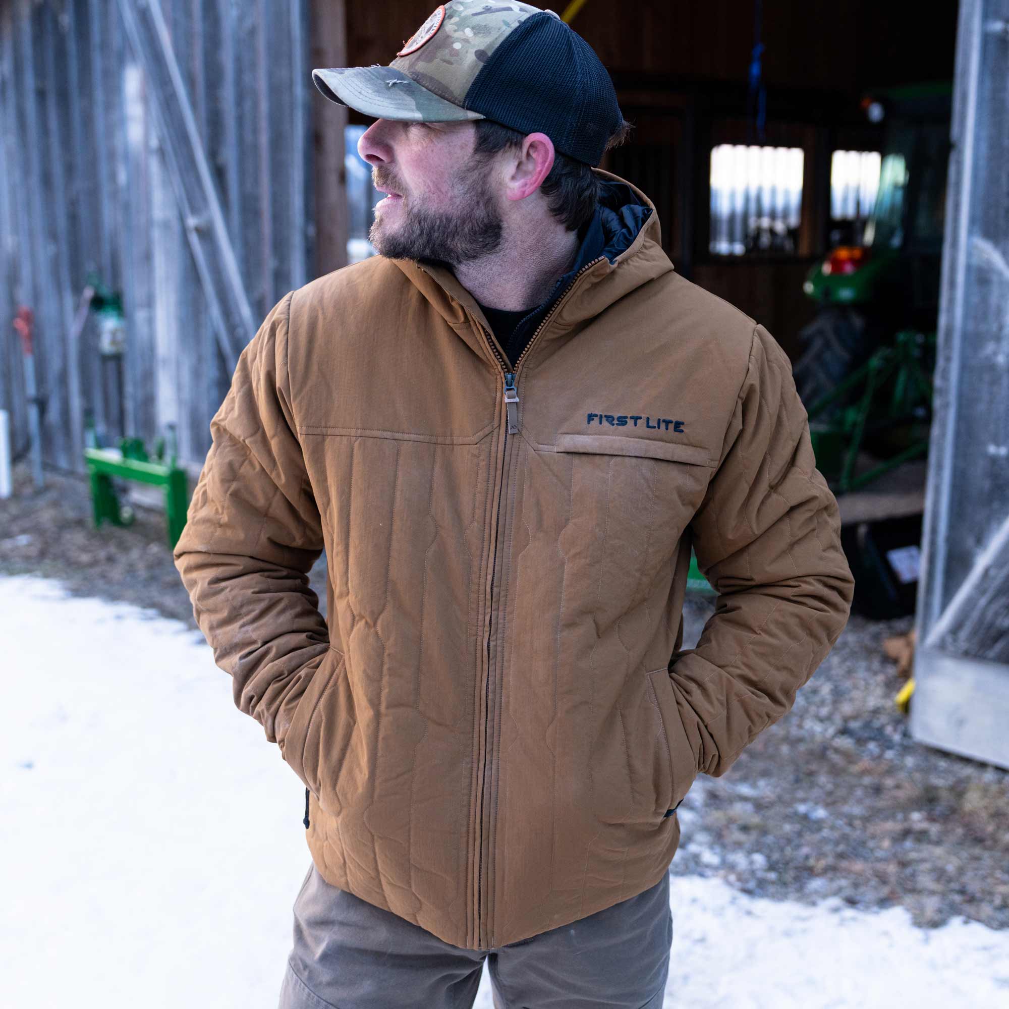 Man wearing a rust outpost jacket with his hands inside the pockets and a camo cap.