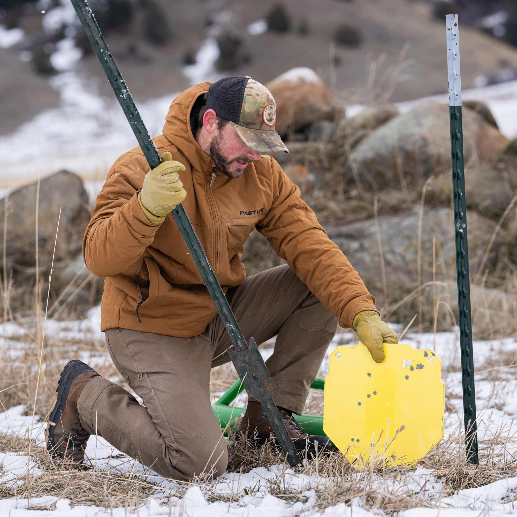 Man on his knee working in the field wearing a rust outpost jacket, yellow gloves, camo cap and brown boots.