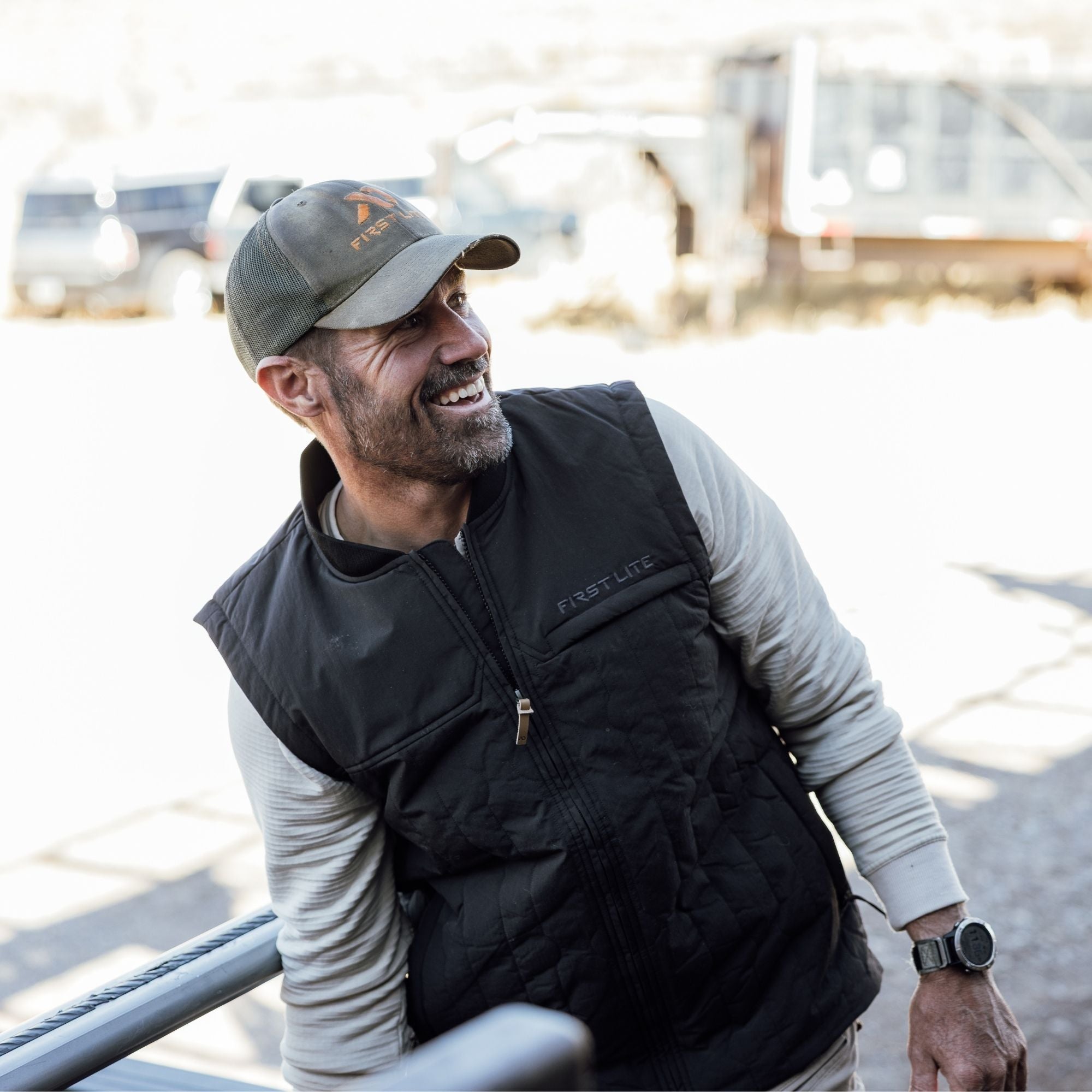 Man wearing a black outpost vest over a light gray shirt and a gray cap with FIRST LITE branding.