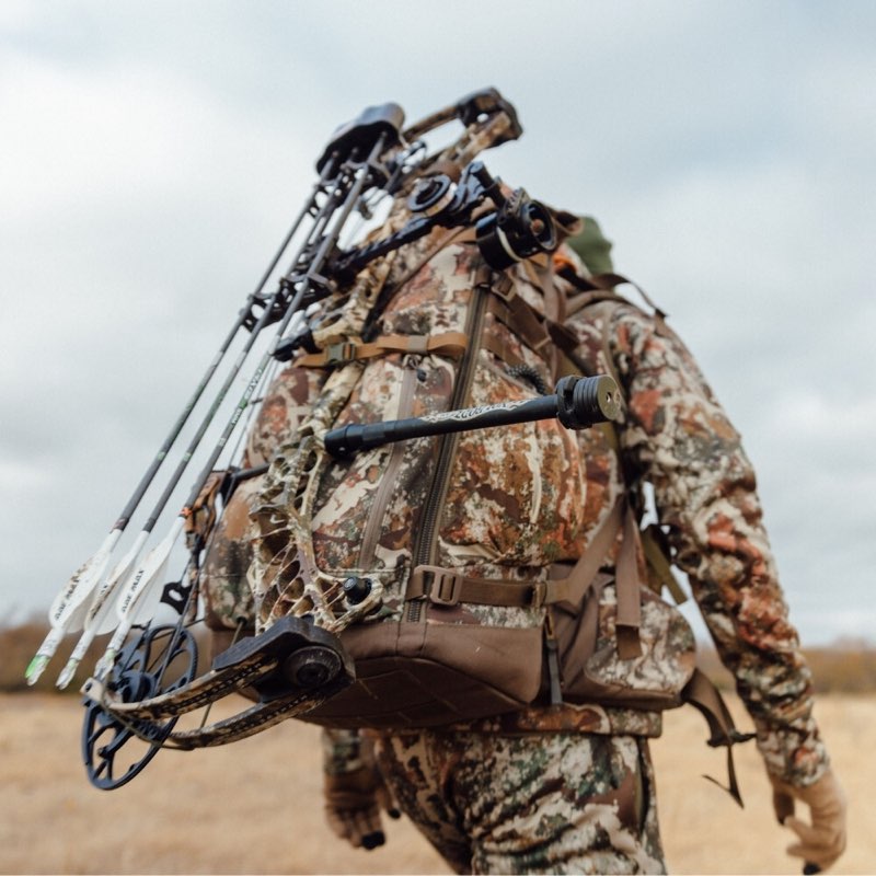 A person in full camouflage outdoor clothing walks through a field while carrying a large backpack loaded with a compound bow and several arrows. The photo is taken from behind, showing the gear in use in an outdoor setting under an overcast sky.