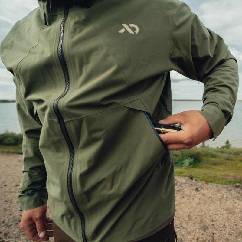 A close-up of a person wearing an olive green waterproof jacket outdoors near water, with a visible white stylized logo on the chest and a hand reaching into a zippered pocket.