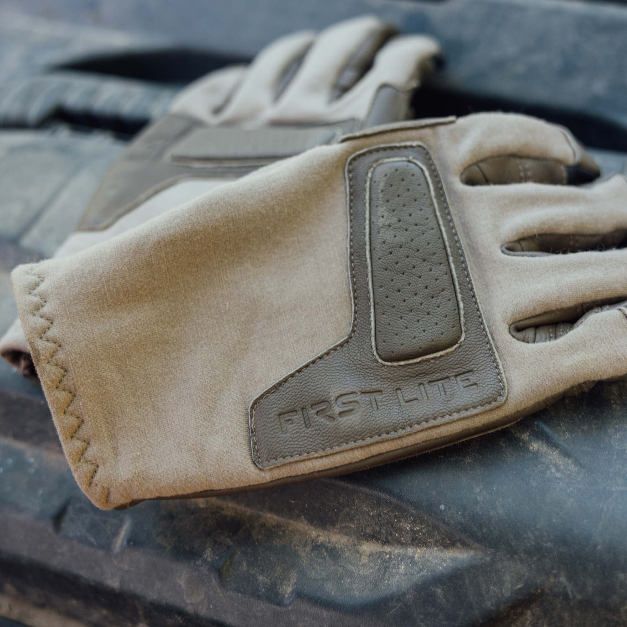 A close-up of a tan leather tactical glove with leather protective panels, including a textured grip panel and an embossed 'FIRST LITE' logo, resting on a black rubber tire.
