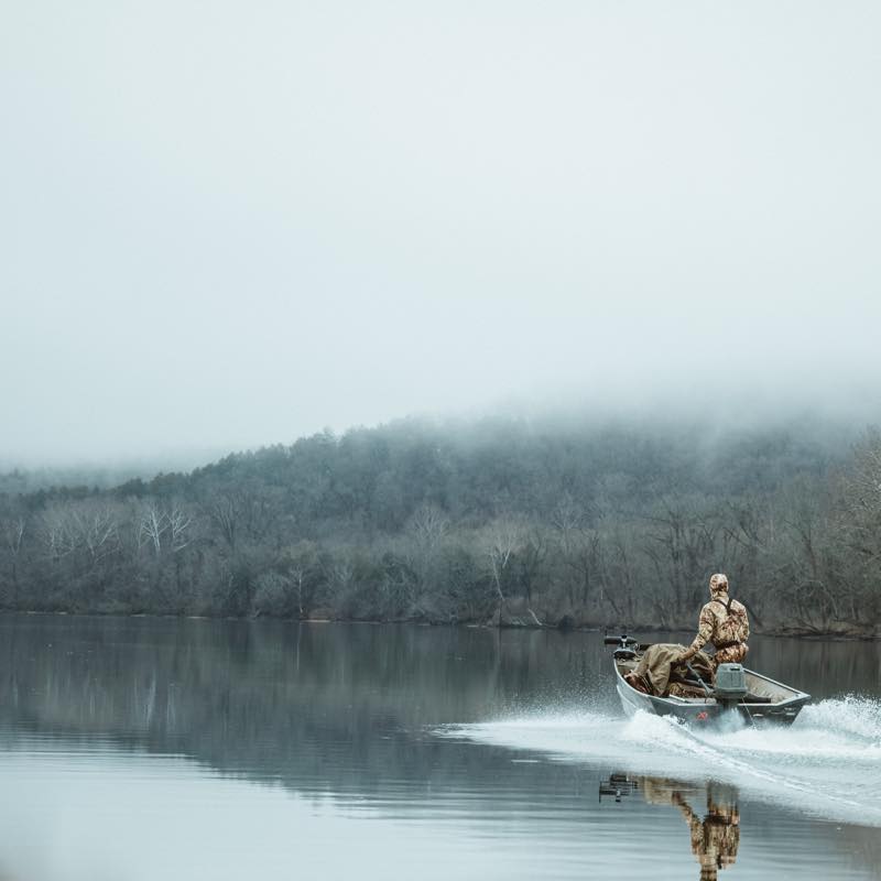 Person in camo drives a green jon boat, creating a wake on a misty lake.