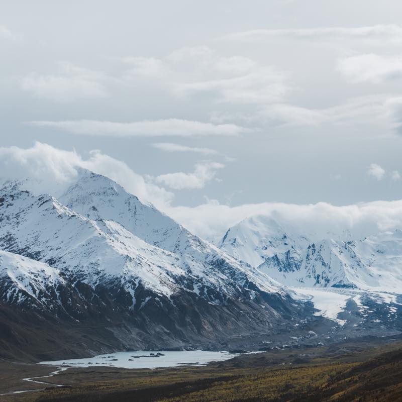 Snow-capped mountains under a cloudy sky, with a glacial lake and winding stream in a valley.