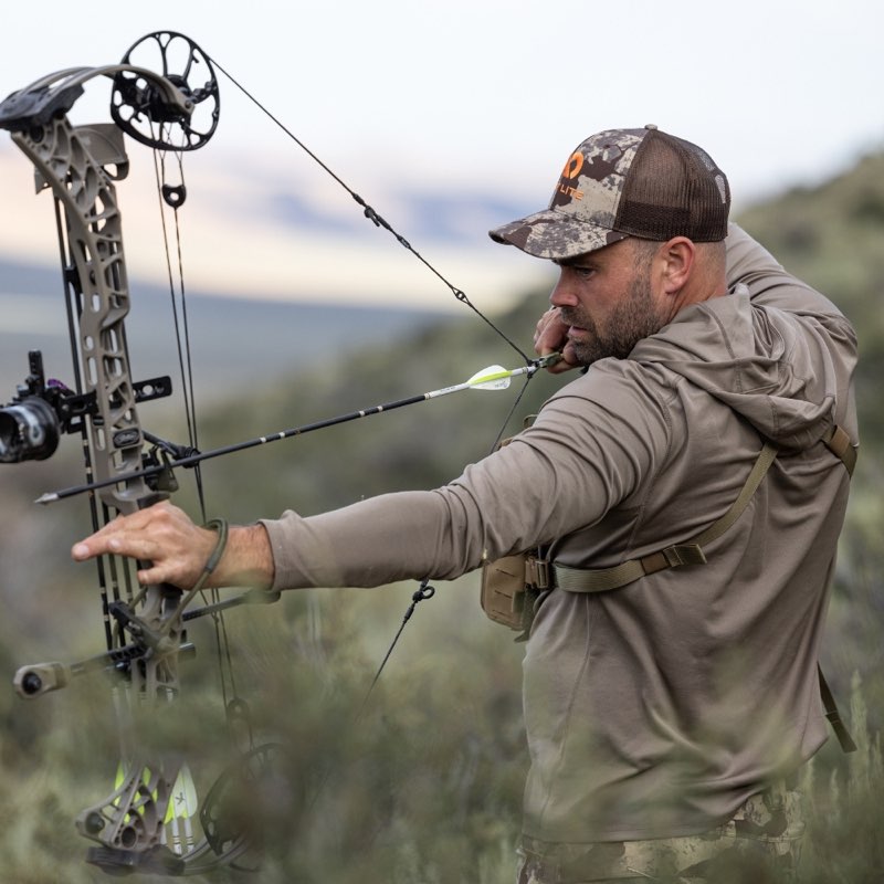 A man in a camouflage cap and tan hunting shirt draws a compound bow in an outdoor field, with arrow nocked. The bow is a detailed gray and black compound bow with visible limbs, pulleys, and sight.