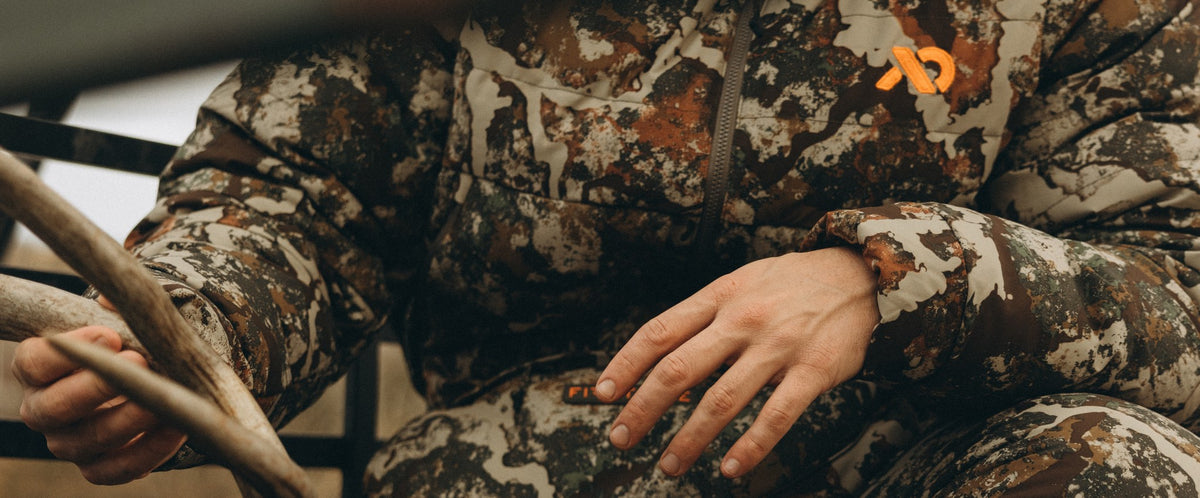 A close-up of a person wearing a camouflage hunting jacket and matching pants, holding an antler. The camouflage pattern features shades of brown, beige, and gray, with a small orange logo on the chest.