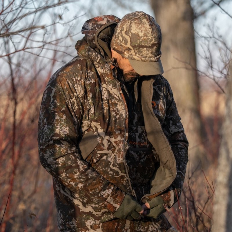 A man wearing a camouflage hunting jacket, matching camouflage cap, and green gloves, standing outdoors in a wooded setting with bare trees and soft warm light.