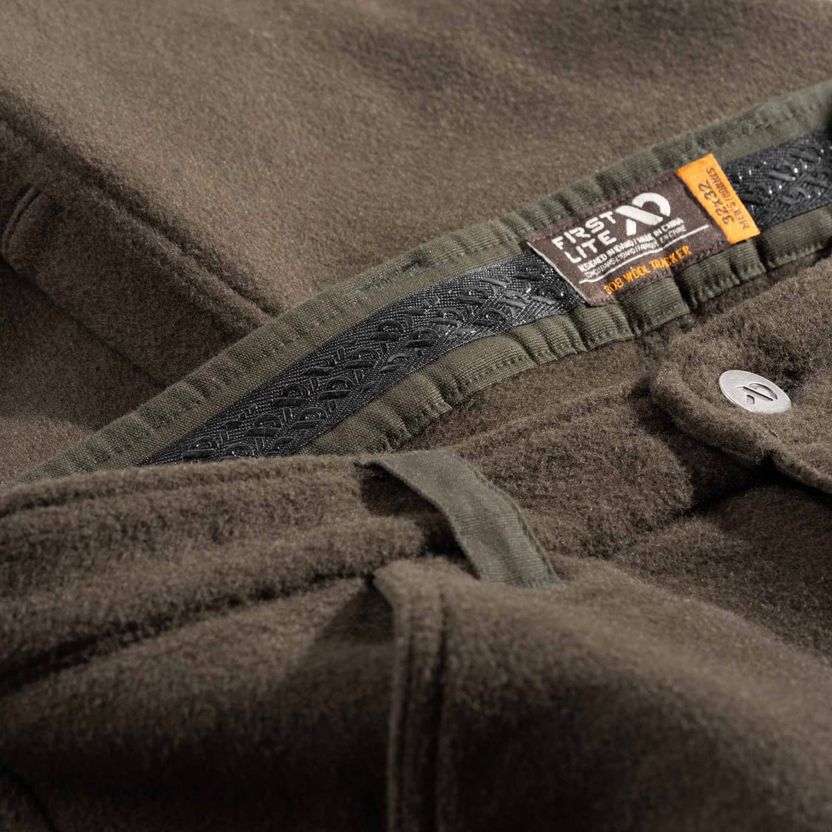 Close-up of a green wool hunting pant showing the inner waistband, a black silicon strip,, and a sewn-in orange and white 'FIRST LITE' brand tag.