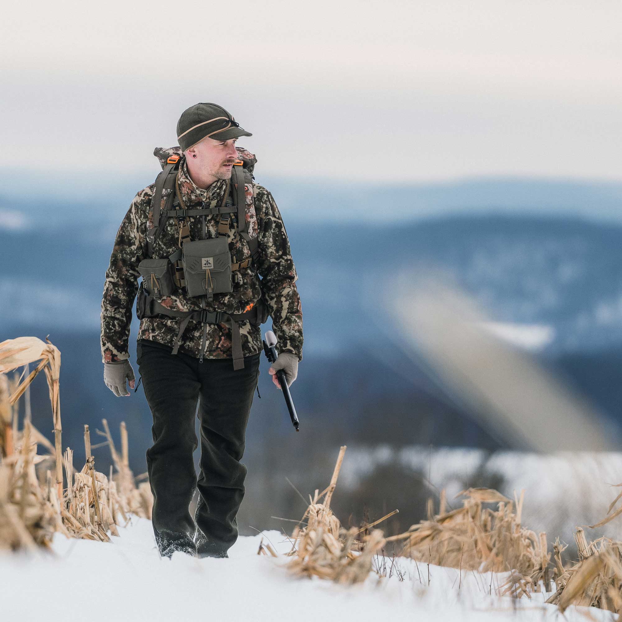 Man walking in the filed wearing camo jacket, dark green cap, light green gloves and dark moss pants.