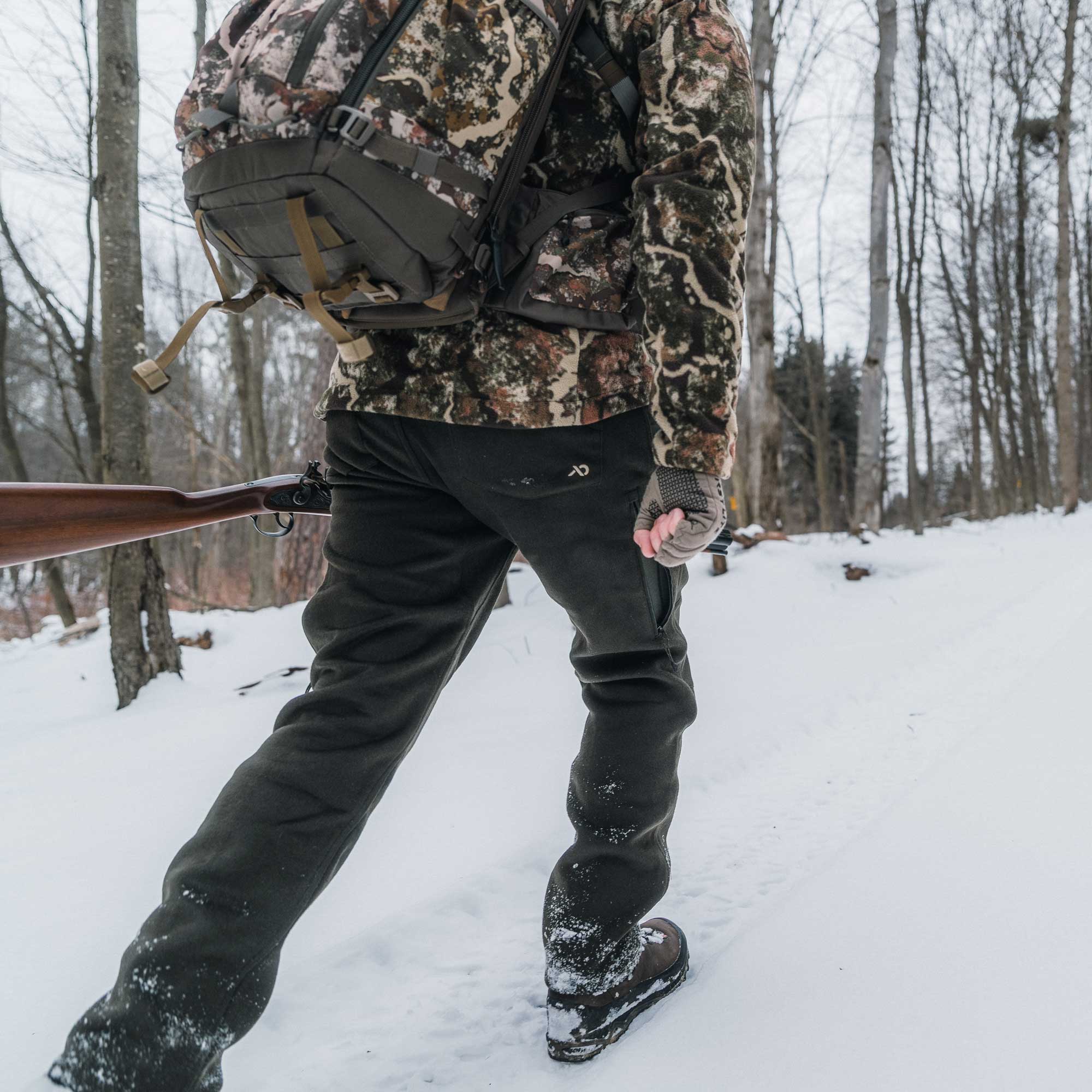 Man walking over snow wearing a camo jacket and dark moss pants, carrying a camo back pack.