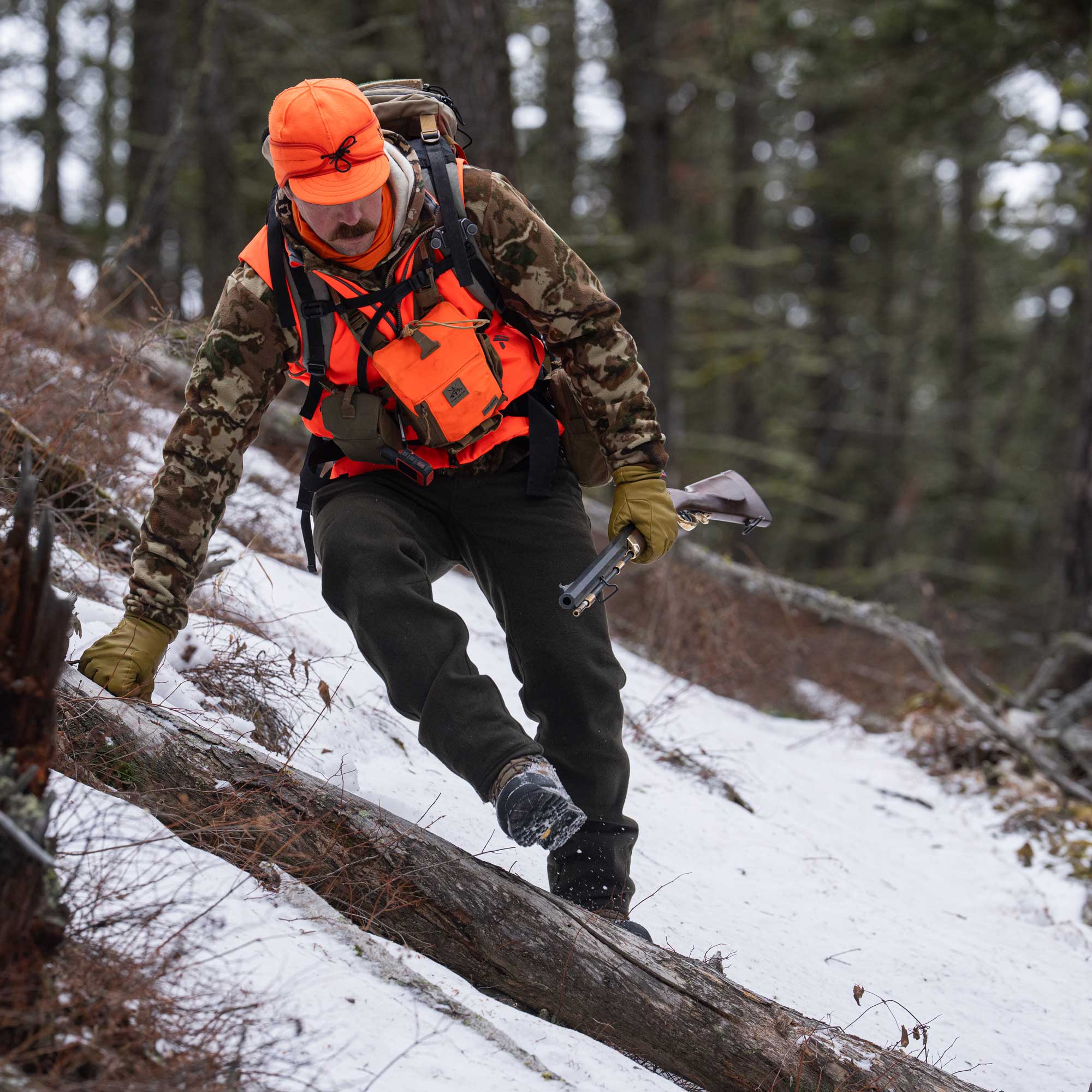 Man in a forest wearing an orange cap and vest over a camo jacket and dark moss pants.
