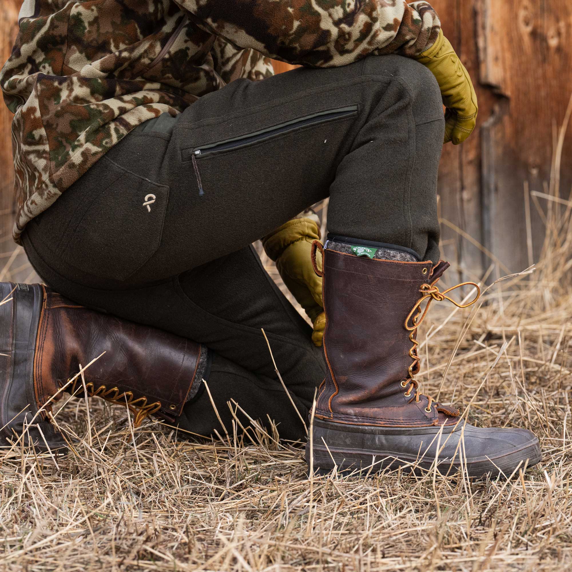 Man with one knee on the floor wearing dark moss pants and high brown boots.